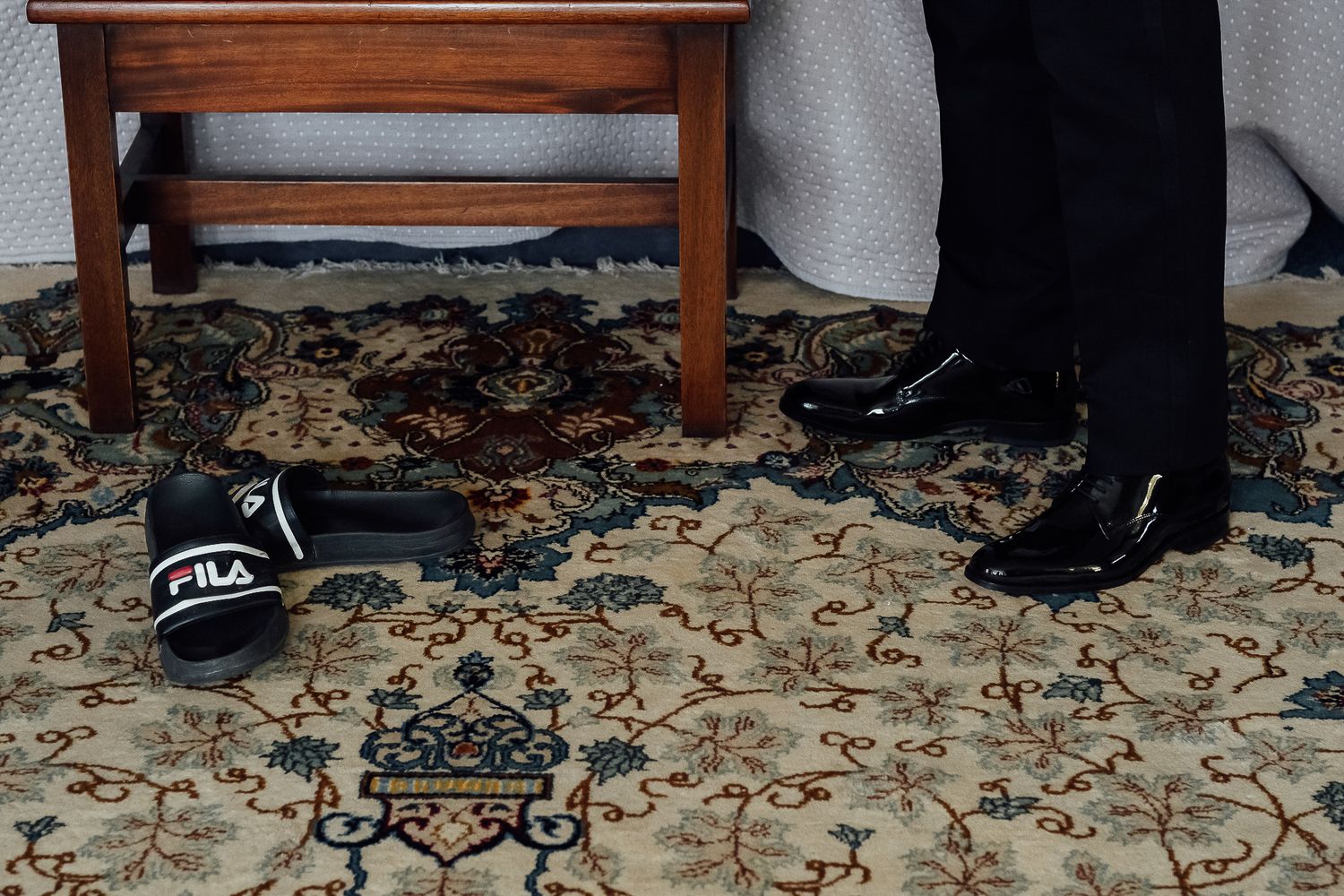Person in black dress shoes standing near a wooden table with FILA slides on a decorative patterned rug.