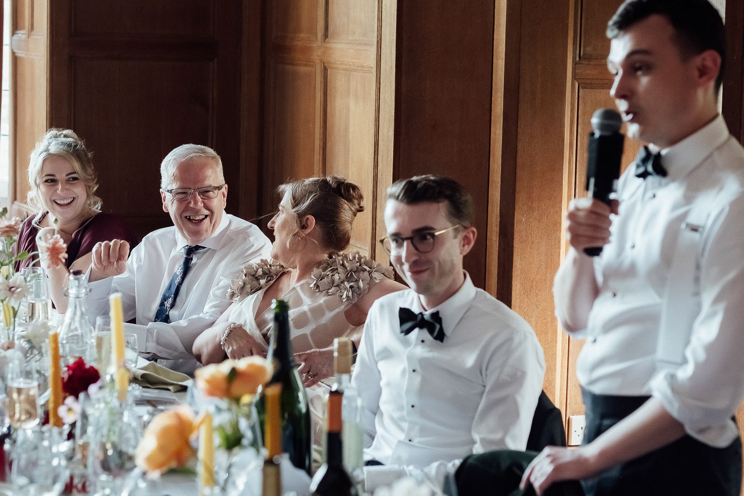 Wedding guests laughing and smiling at a reception dinner table during a heartfelt speech at an elegant venue.