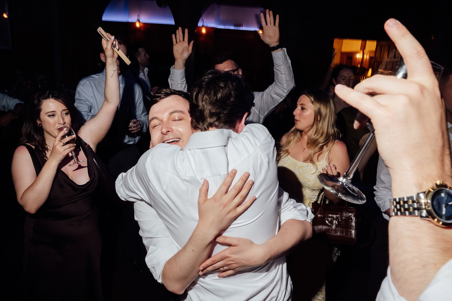 Two men share a joyful hug on a lively wedding dance floor surrounded by celebrating guests.