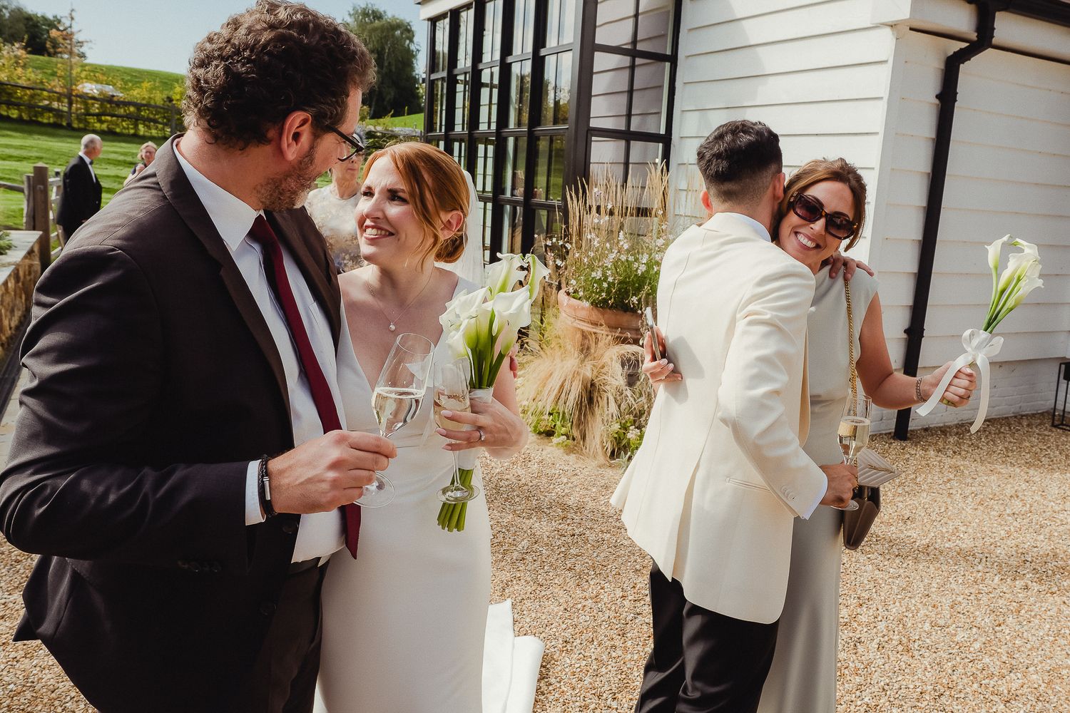 Wedding guests mingle and chat while holding champagne glasses on a sunny patio outside a white building.
