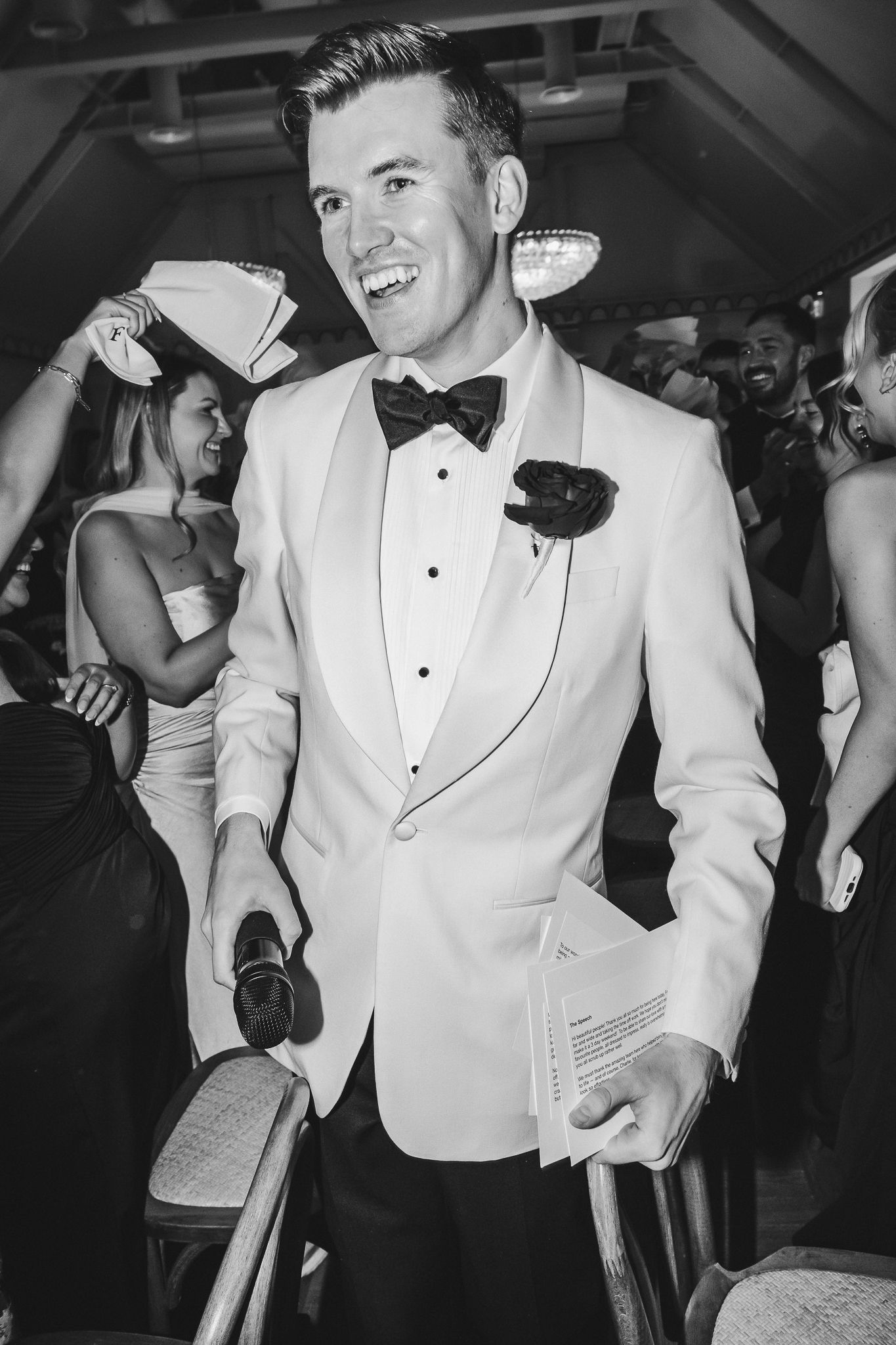 A groom in a white tuxedo jacket and black bow tie dances and smiles during a wedding reception.