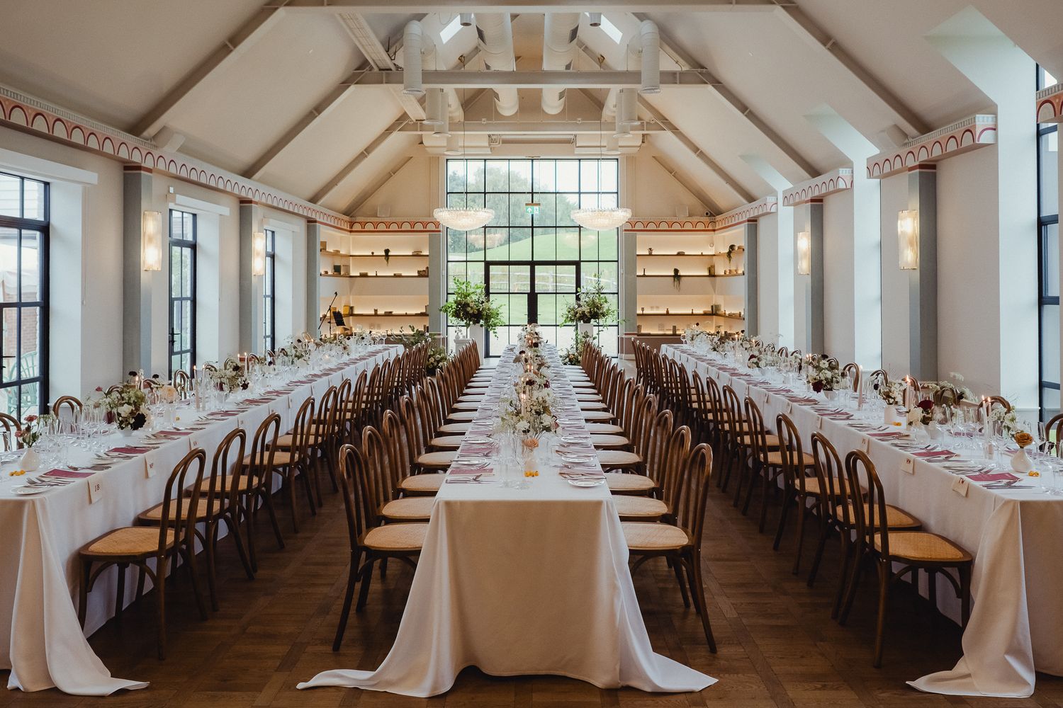 Long banquet tables with white linens and chairs set up in an elegant reception hall with natural light streaming through windows.