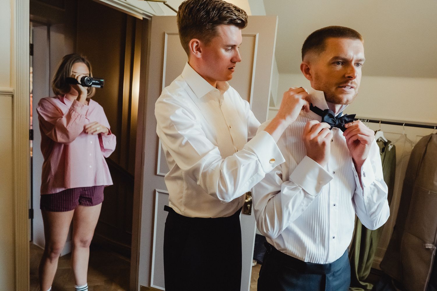 Wedding party members help each other adjust bowties and formal attire while getting ready in a hallway.