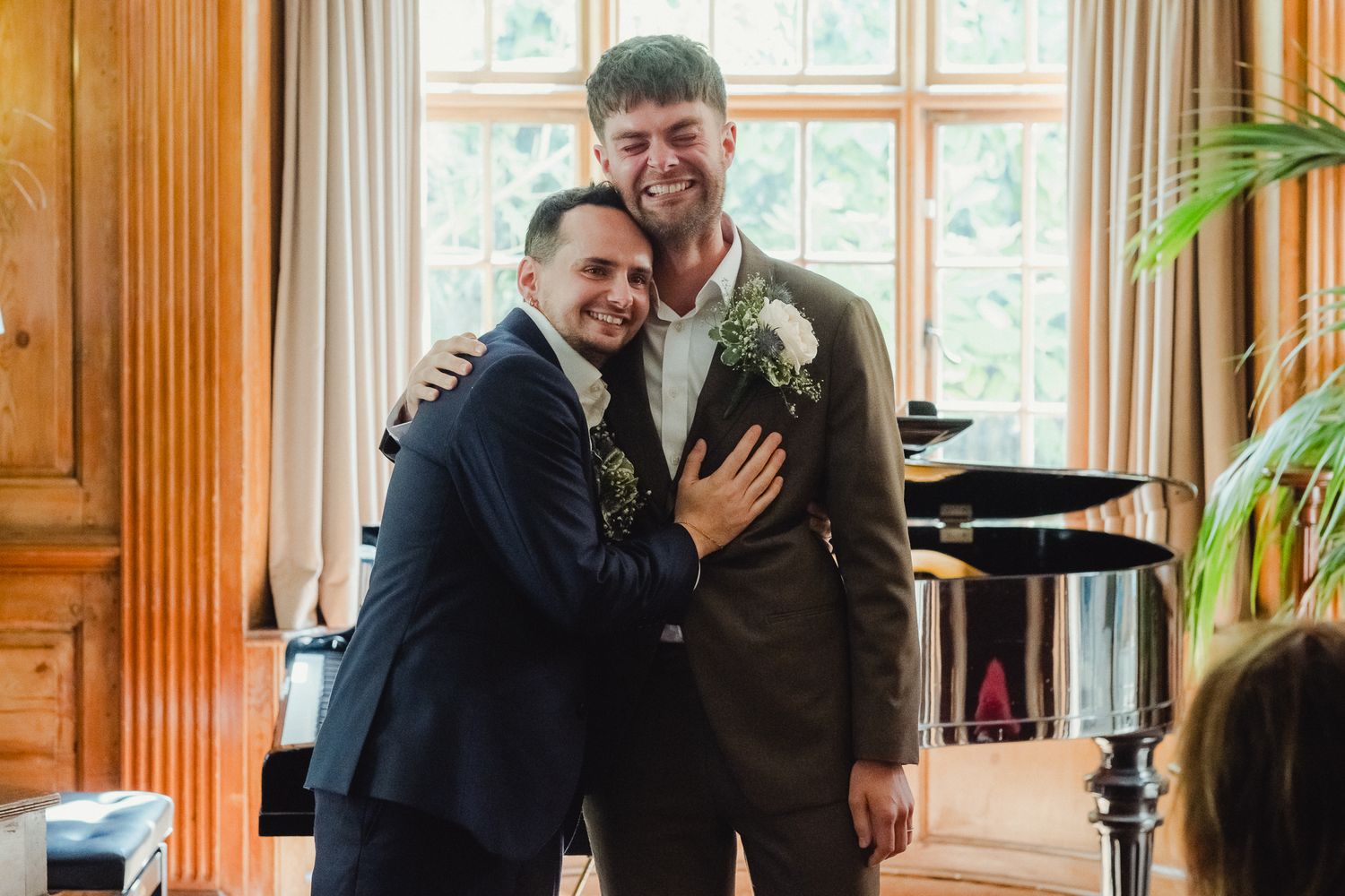 Two people in formal suits share a happy moment together in an elegant room with wood paneling and windows.