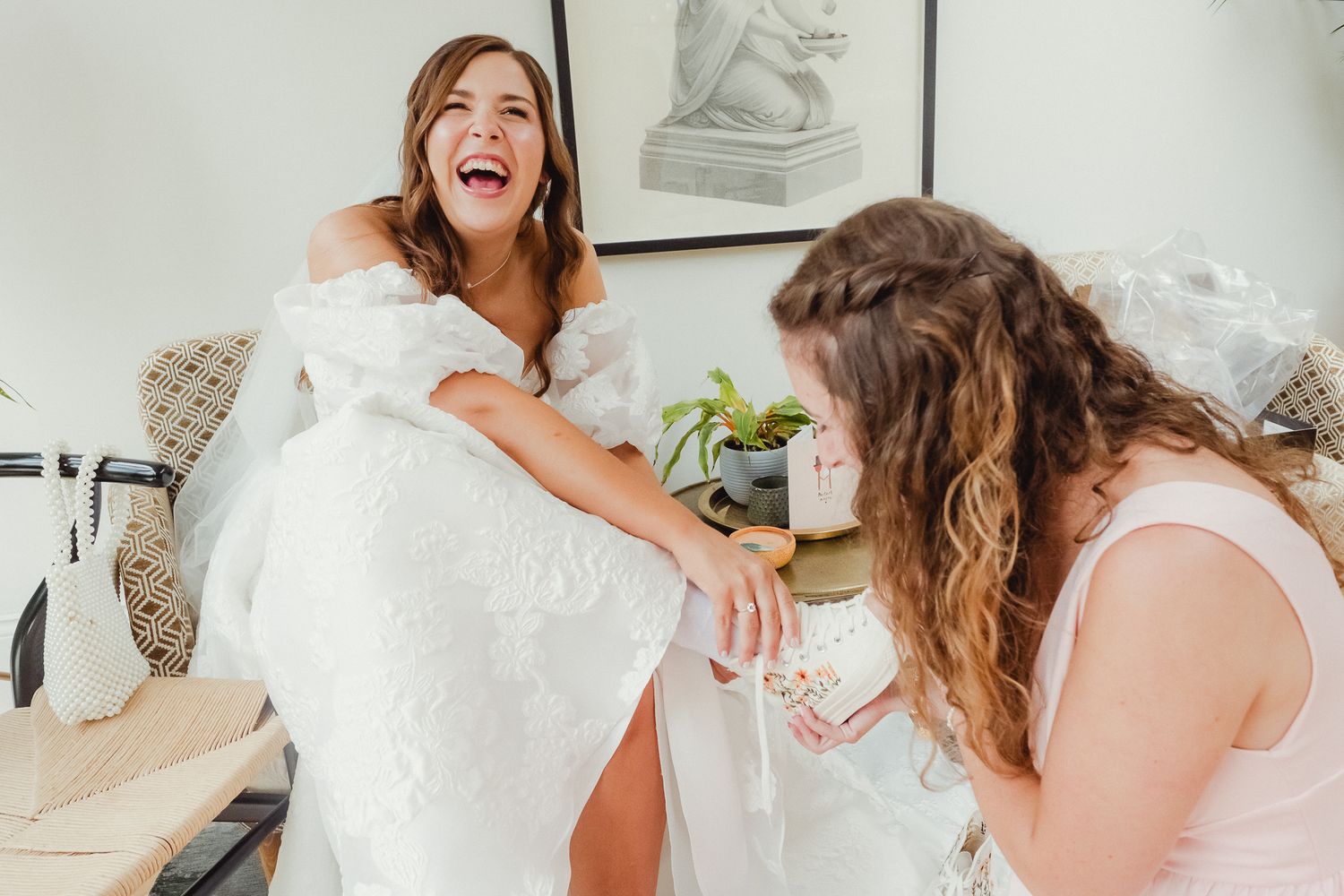 Two people share a joyful moment while preparing for a wedding with a white dress in a bright indoor setting.