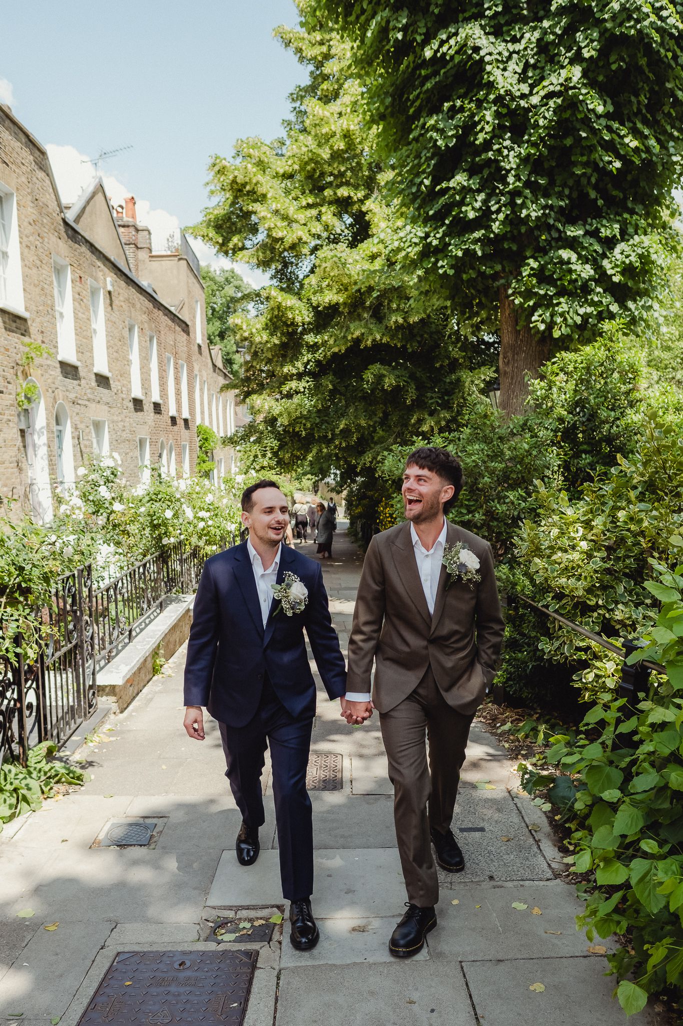 Two people in suits walk and laugh together down a tree-lined residential street with brick row houses.
