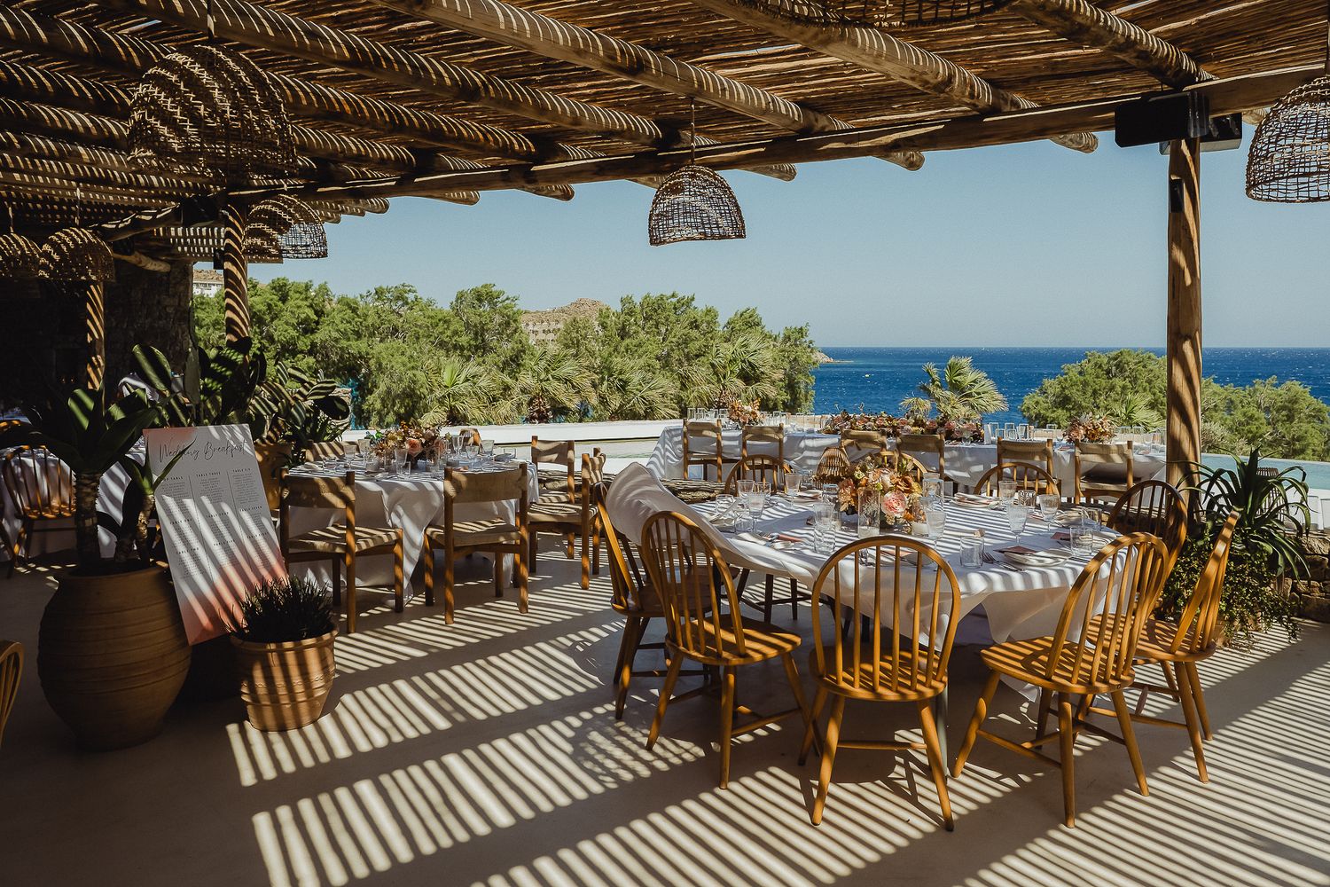 Wooden chairs and tables under a rustic beach pergola overlooking the Mediterranean Sea on a sunny summer day.