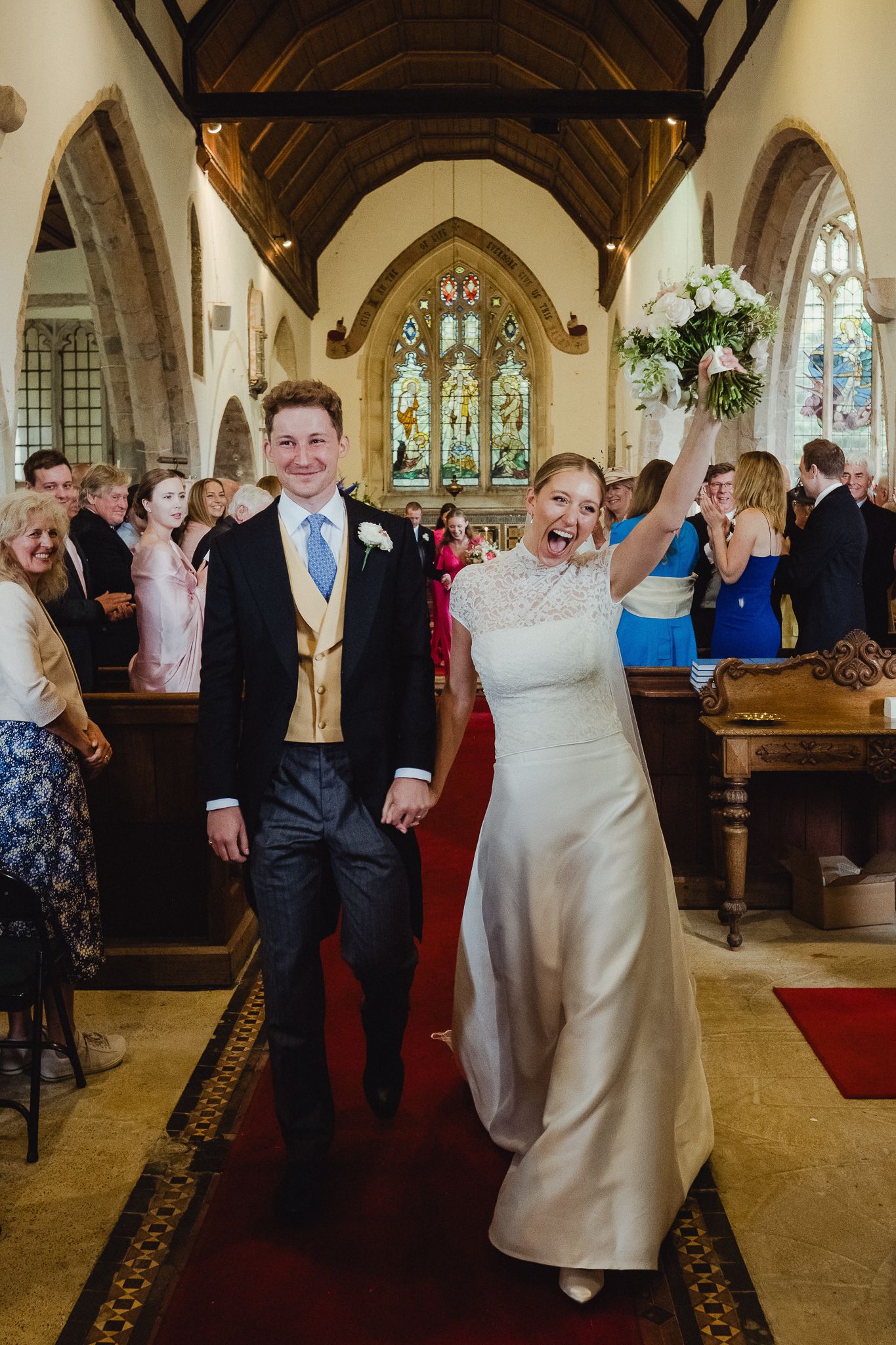 Wedding guests dance joyfully in the church hall during a reception celebration with stained glass windows in background.