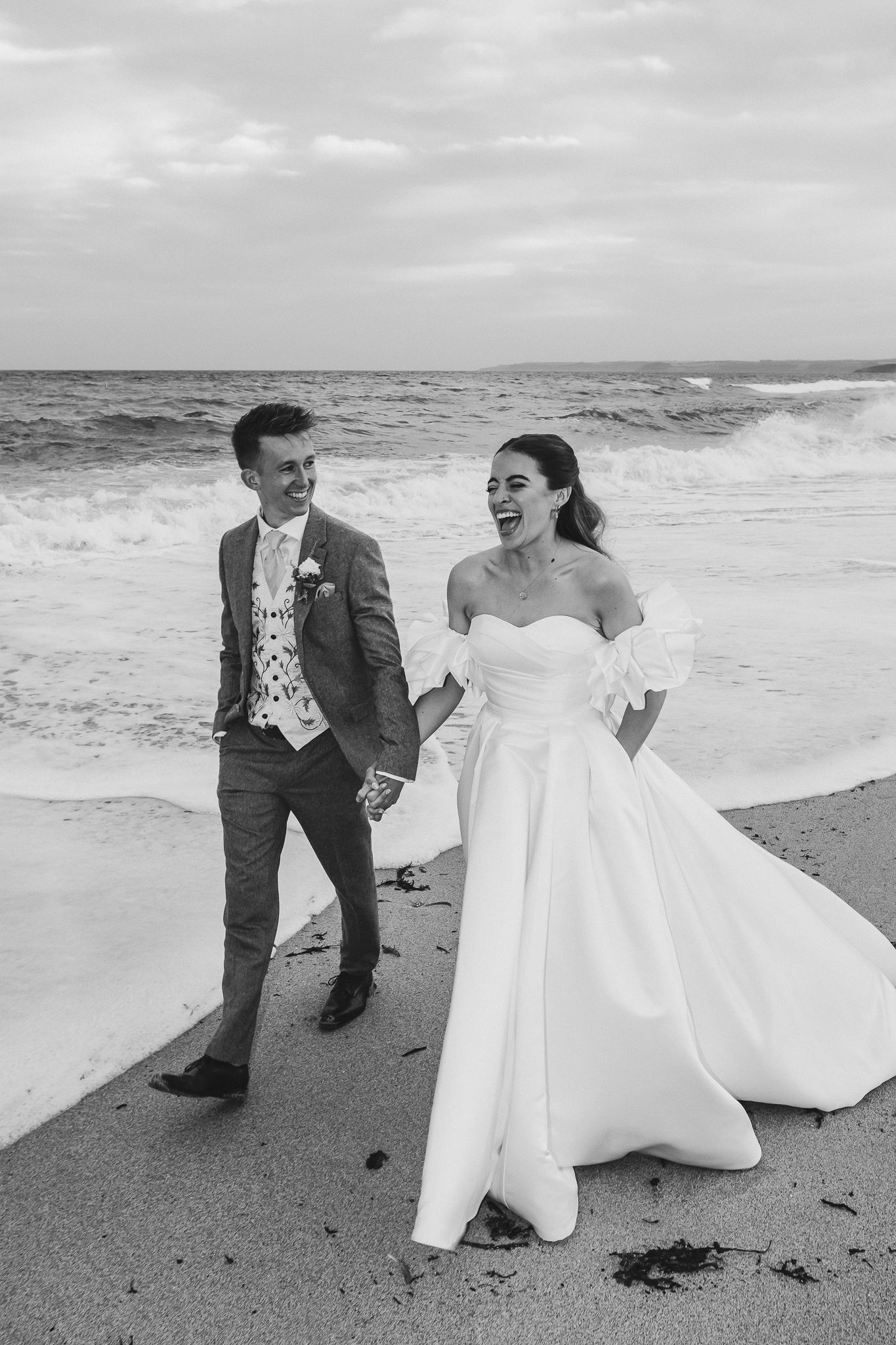 A joyful couple in wedding attire shares candid moments on a cloudy beach in black and white photography.