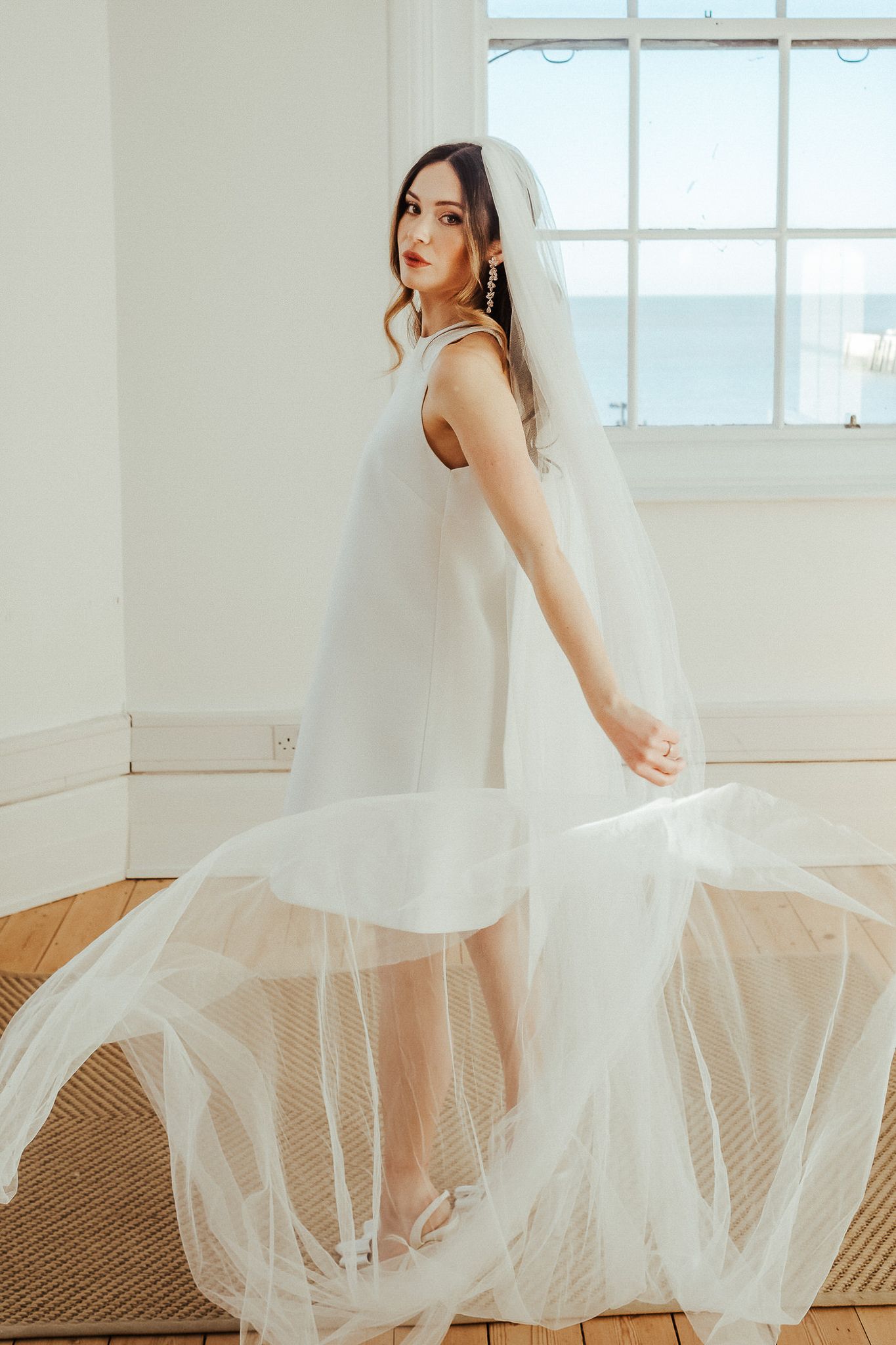 A bride in a flowing white tulle gown twirls gracefully in a bright sunlit room with large windows.