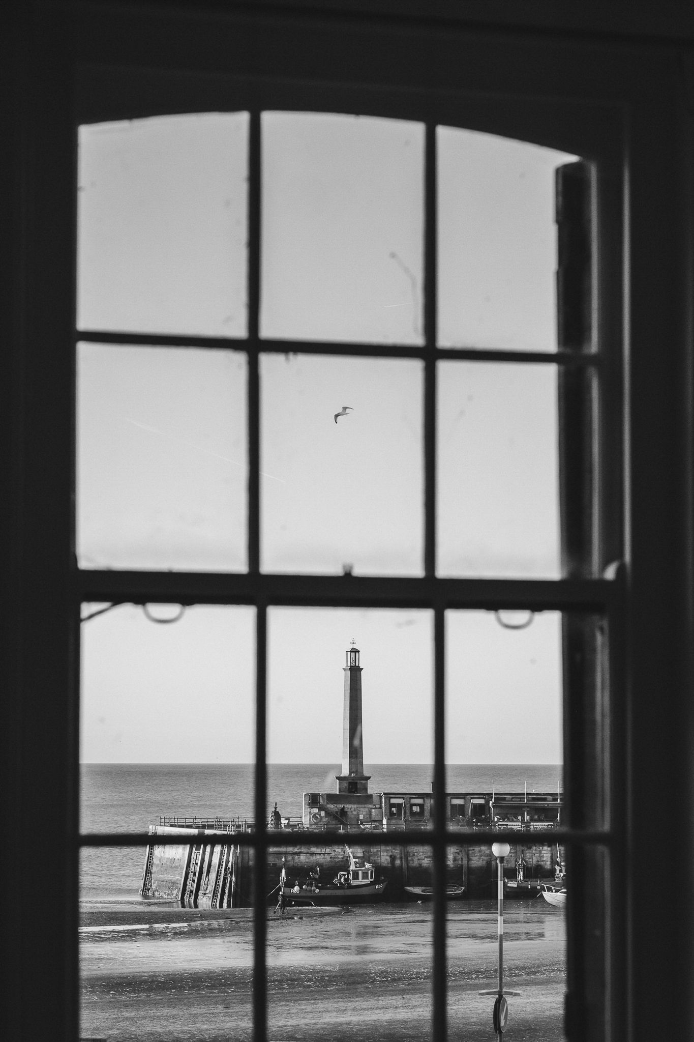 Black and white view through window frames showing a lighthouse and pier on the ocean horizon.