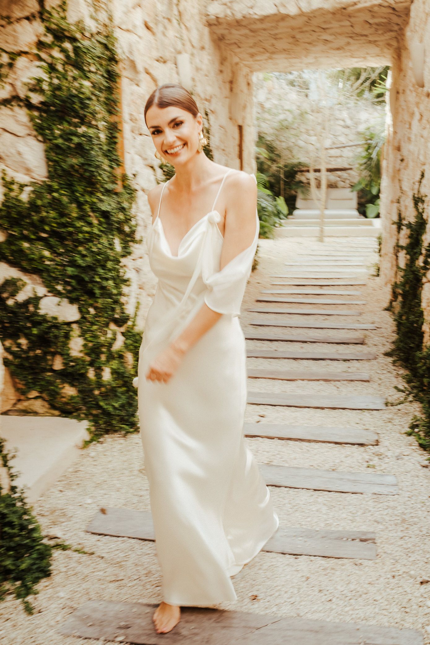 A person in a flowing white dress runs joyfully up stone steps surrounded by greenery and sunlight.