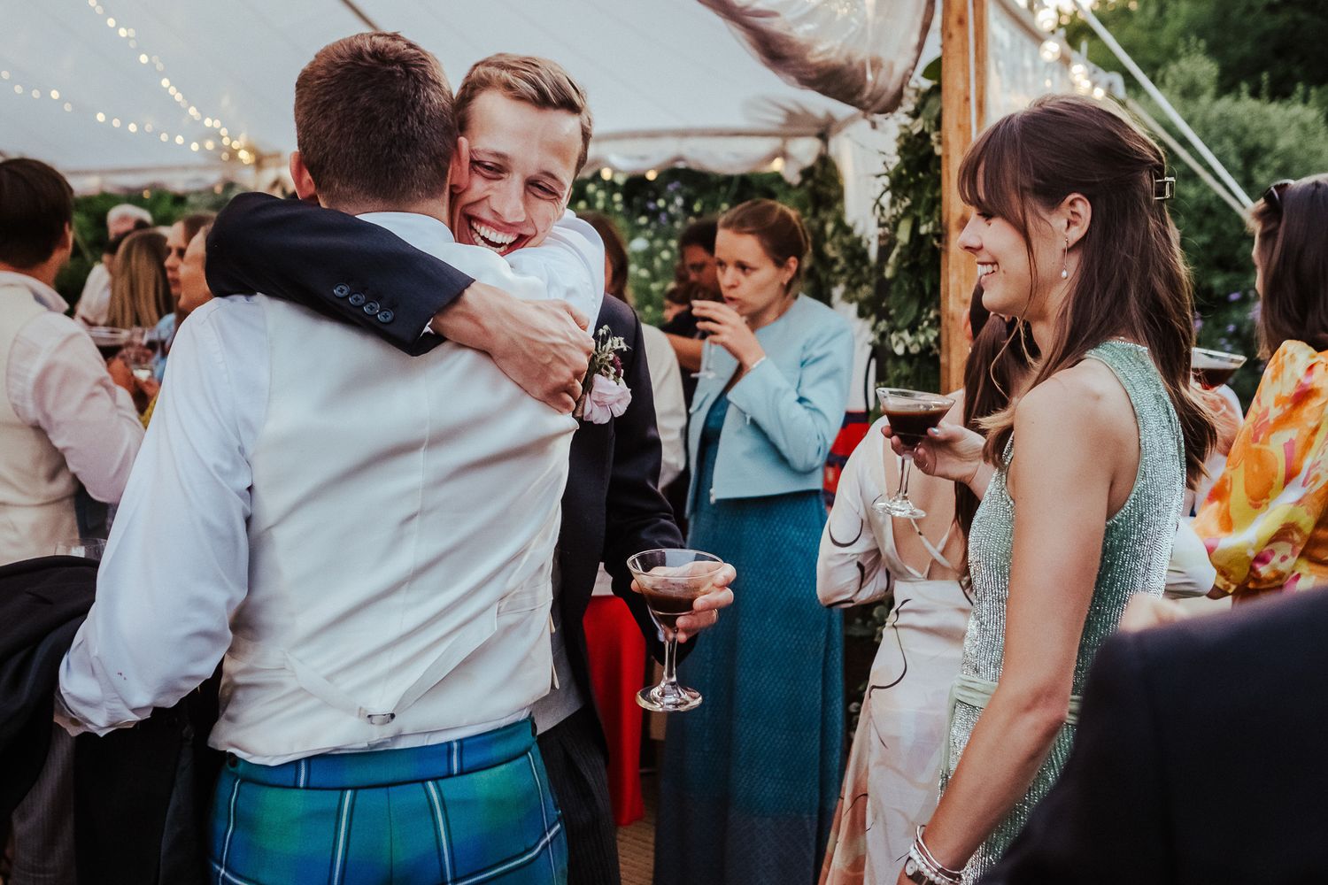 Wedding guests share hugs and celebrations under a decorated tent at an outdoor reception.