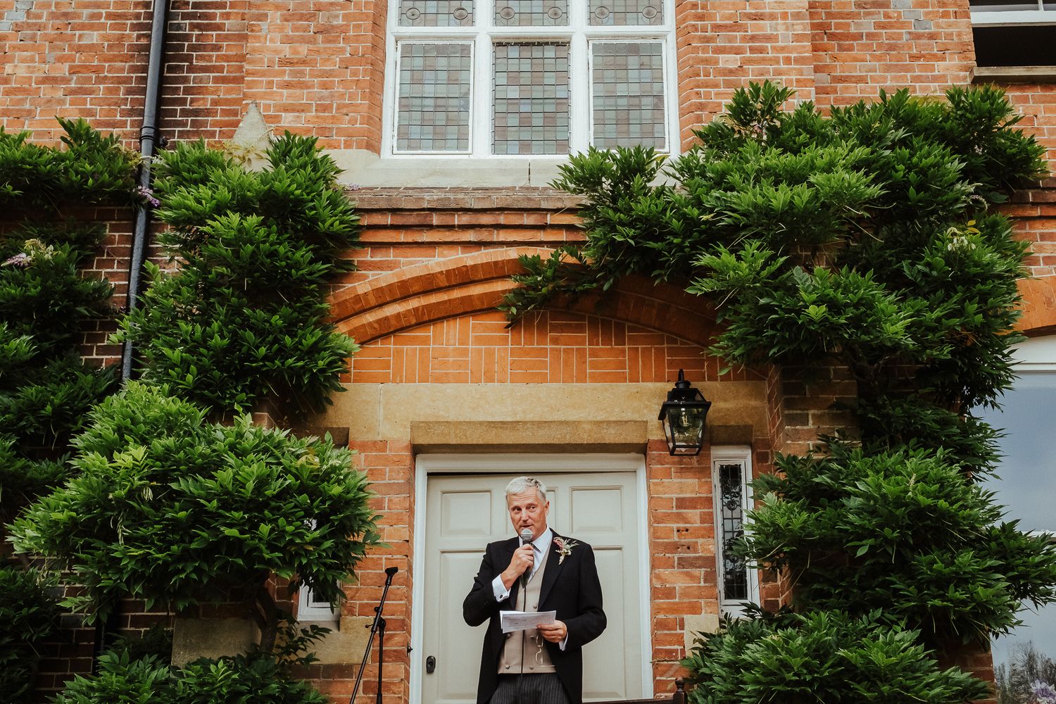 A figure in formal attire stands in front of a brick building entrance with greenery.