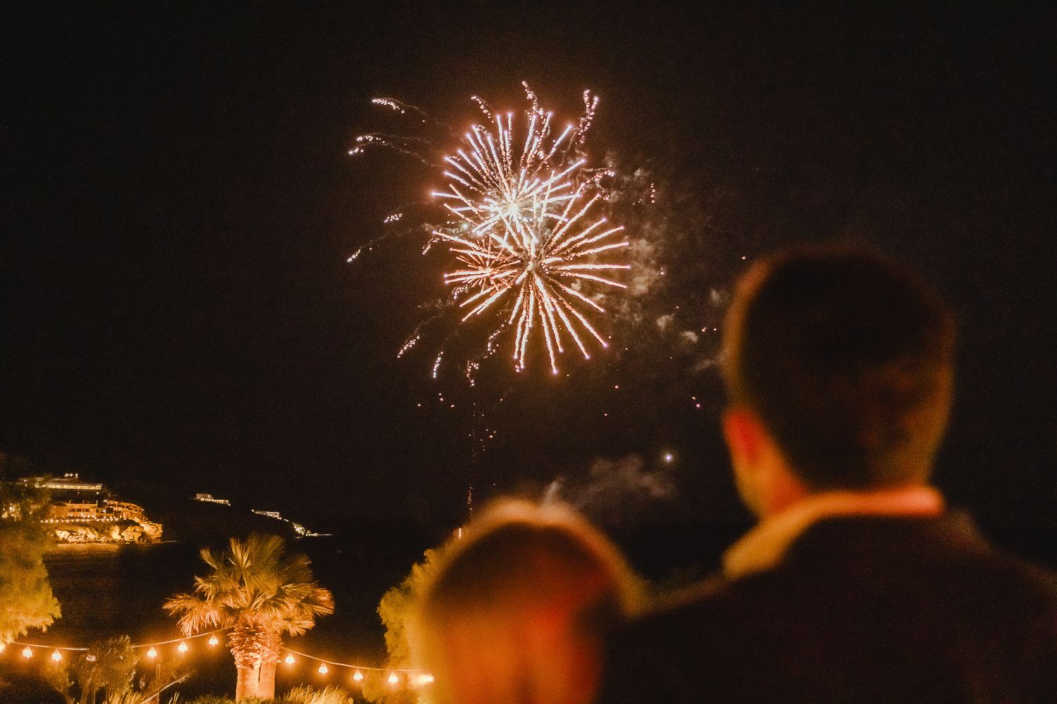 People watching bright fireworks display bursting in night sky above illuminated buildings at evening celebration.