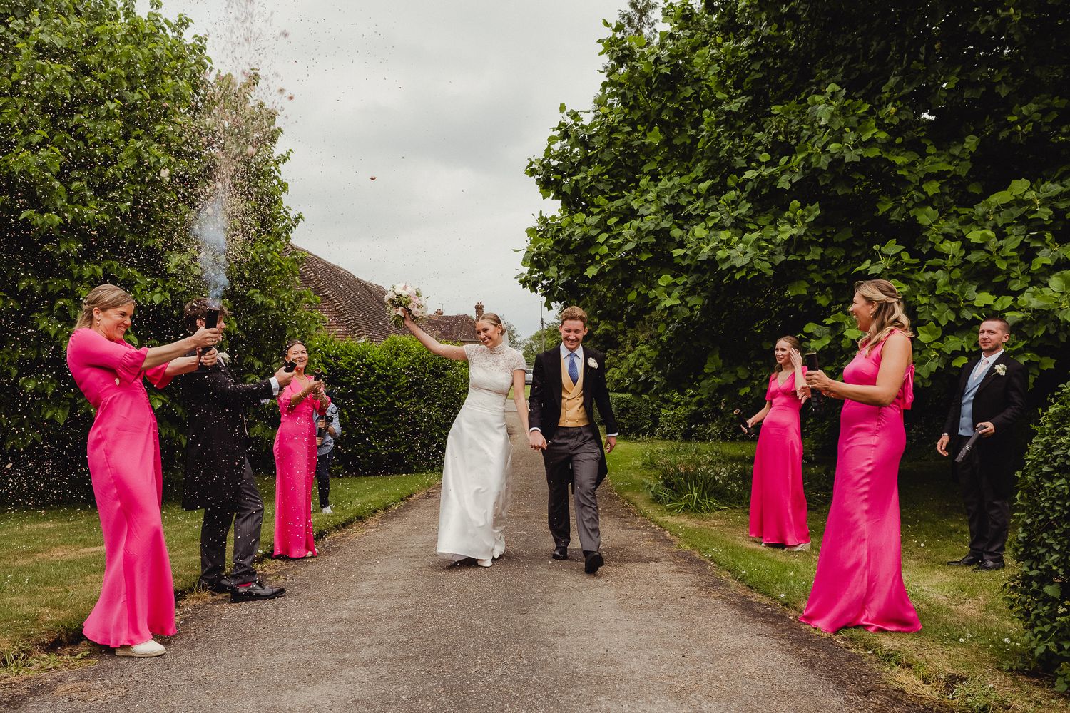 Wedding party members in bright pink dresses pose for photos with newlyweds along a scenic tree-lined path.