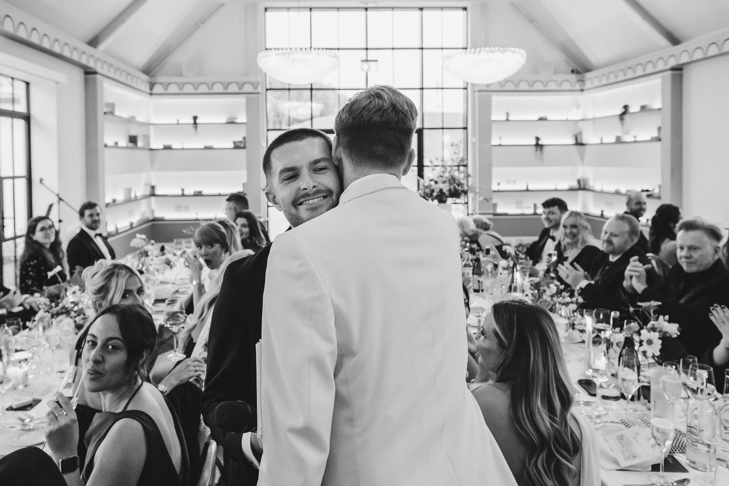 Black and white photo of a lively wedding reception with guests seated at tables in a bright room with white walls.