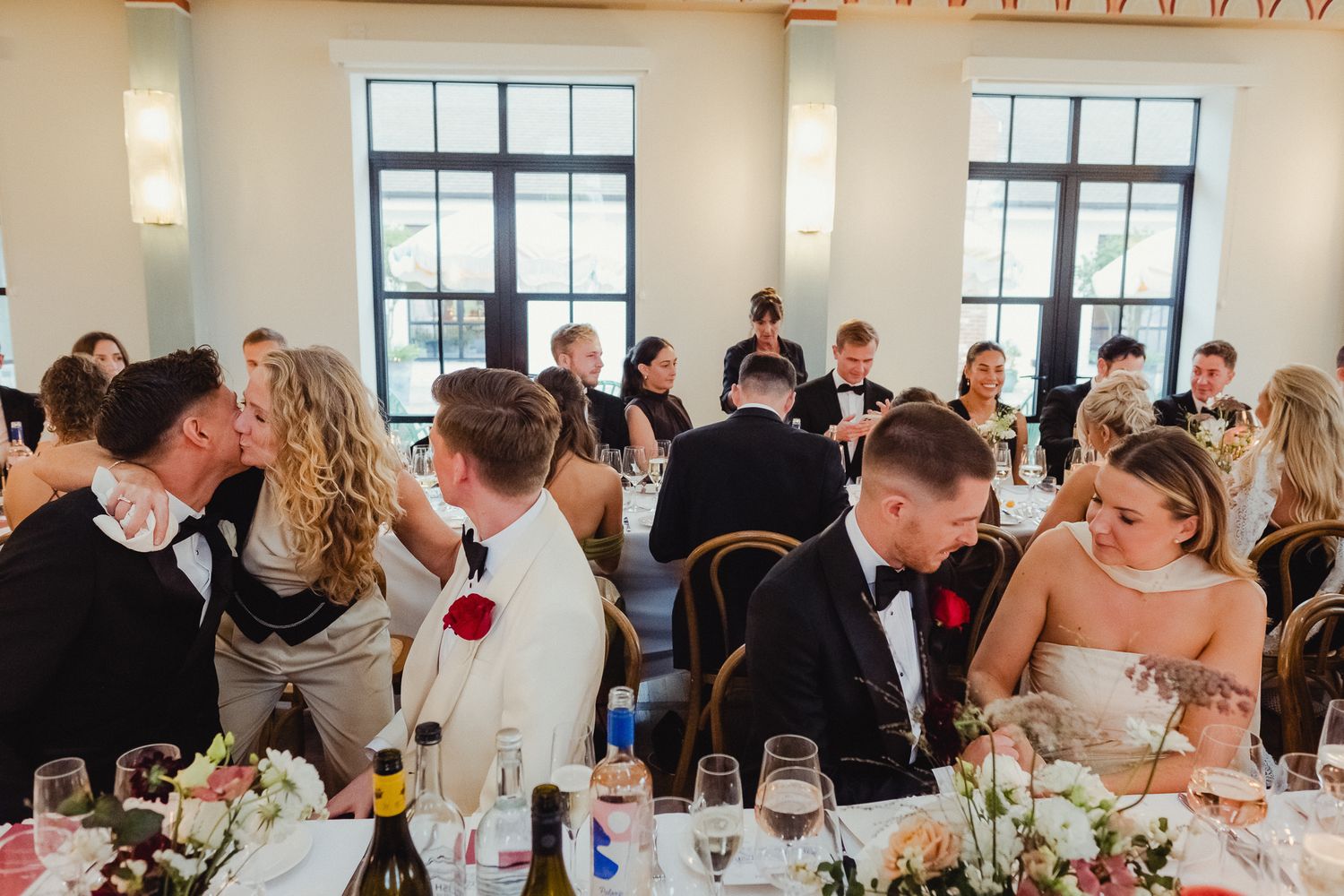 Wedding reception guests seated at long tables in an elegant banquet hall with natural lighting and floral centerpieces.