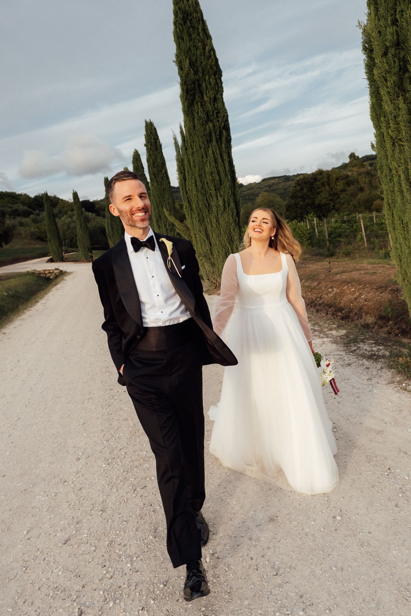 Bride in a flowing white gown and groom in a black tuxedo walk hand in hand along a gravel road lined with tall cypress trees.