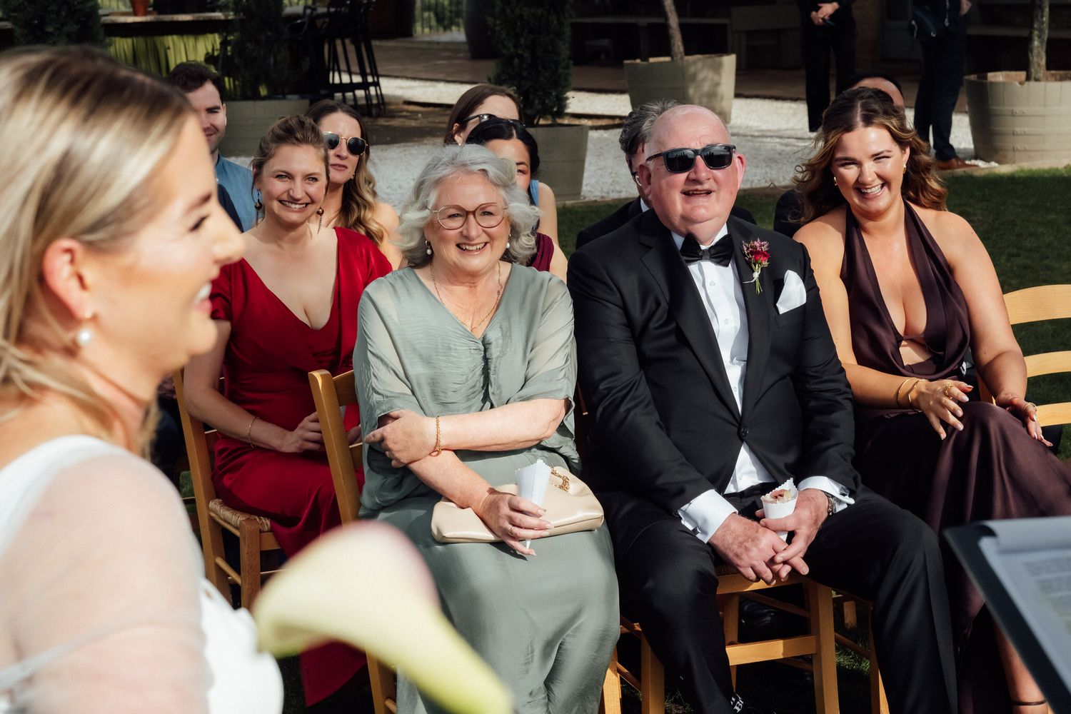 Smiling wedding guests in formal attire sit on wooden chairs outdoors, watching a ceremony with joyful expressions.