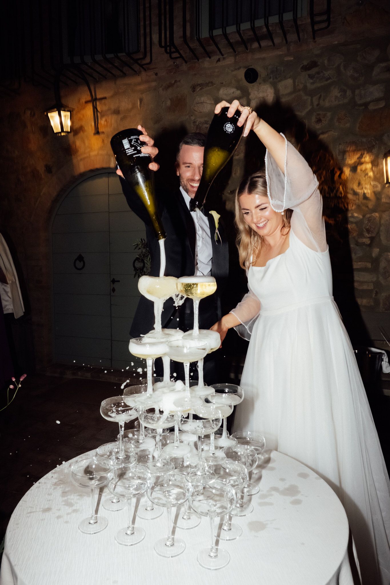 Bride and groom pour champagne from two bottles over a tiered champagne tower at a candlelit stone courtyard wedding reception.