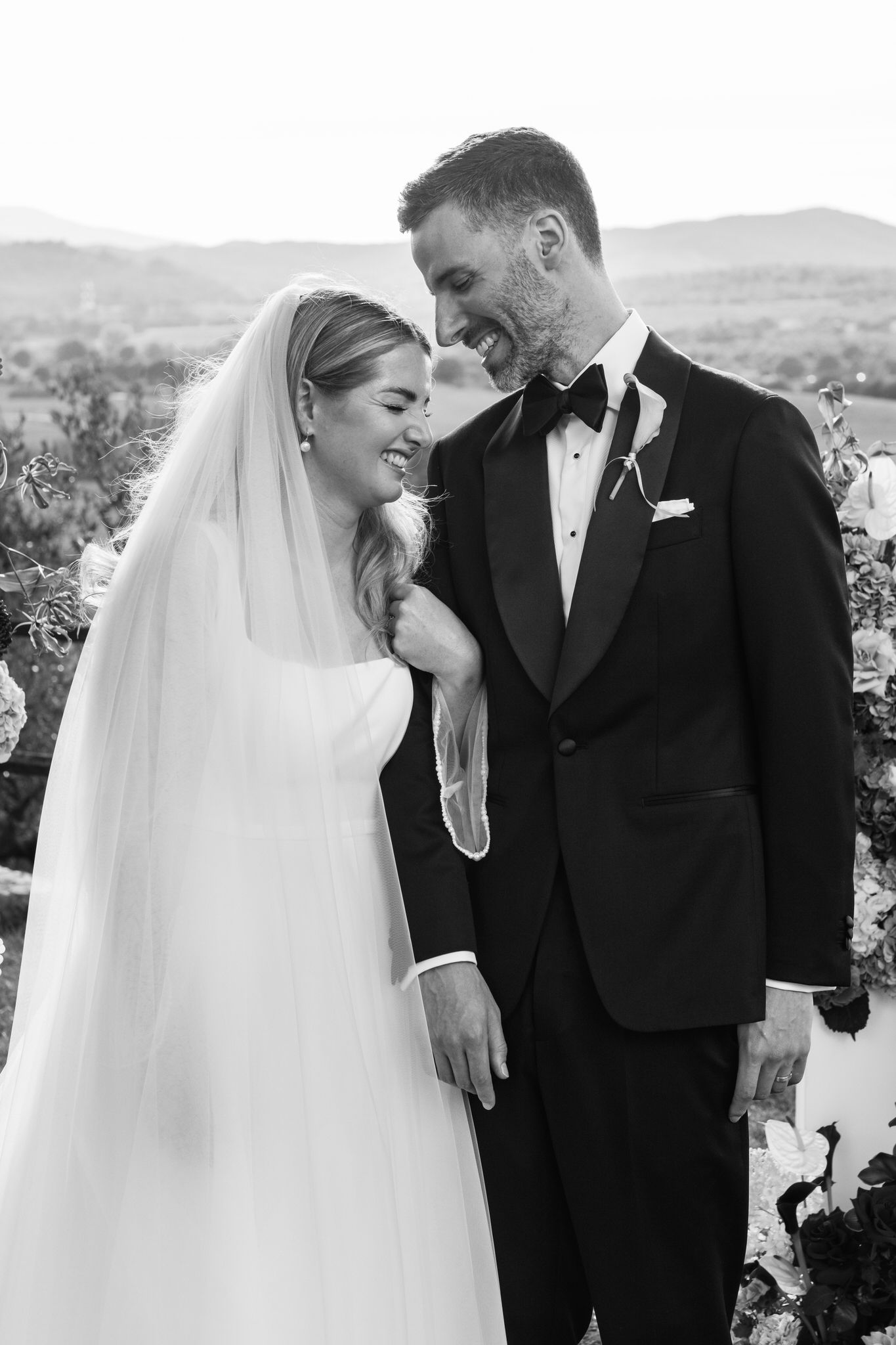 Black and white photo of a laughing bride in a long veil embracing a smiling groom in a tuxedo and bow tie, with hills behind them.