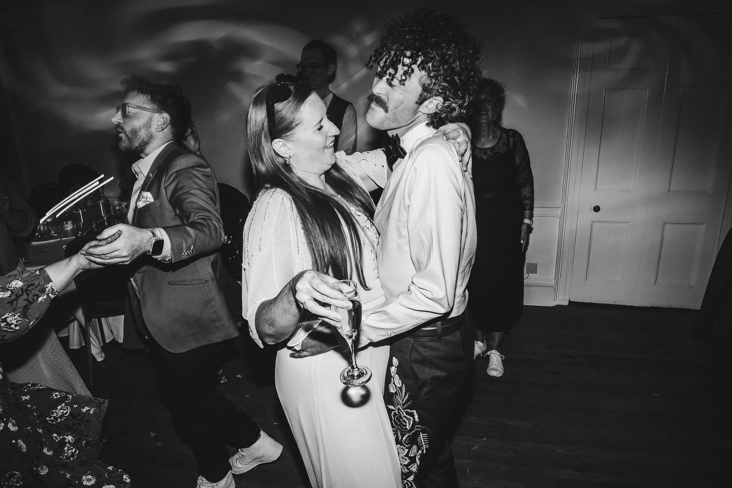 Black and white photo of a laughing couple dancing closely at a lively wedding reception while guests dance behind them.