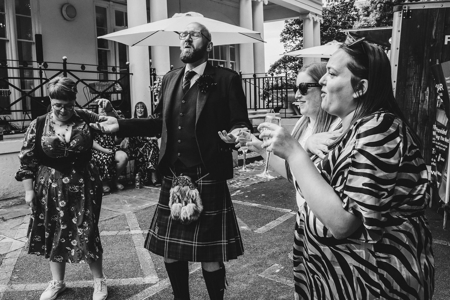 Bearded man in a kilt and sporran gestures while chatting with wedding guests outside beneath a white parasol.