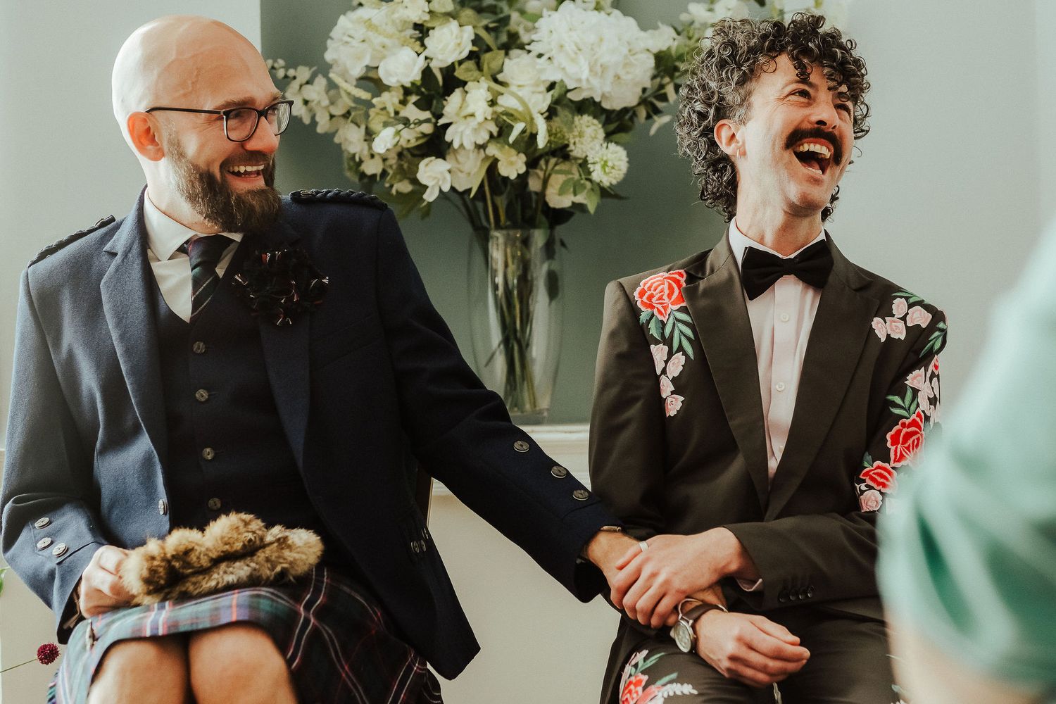 Two grooms hold hands and laugh during their wedding ceremony, one in a tartan kilt outfit and the other in a floral blazer with a bow tie.