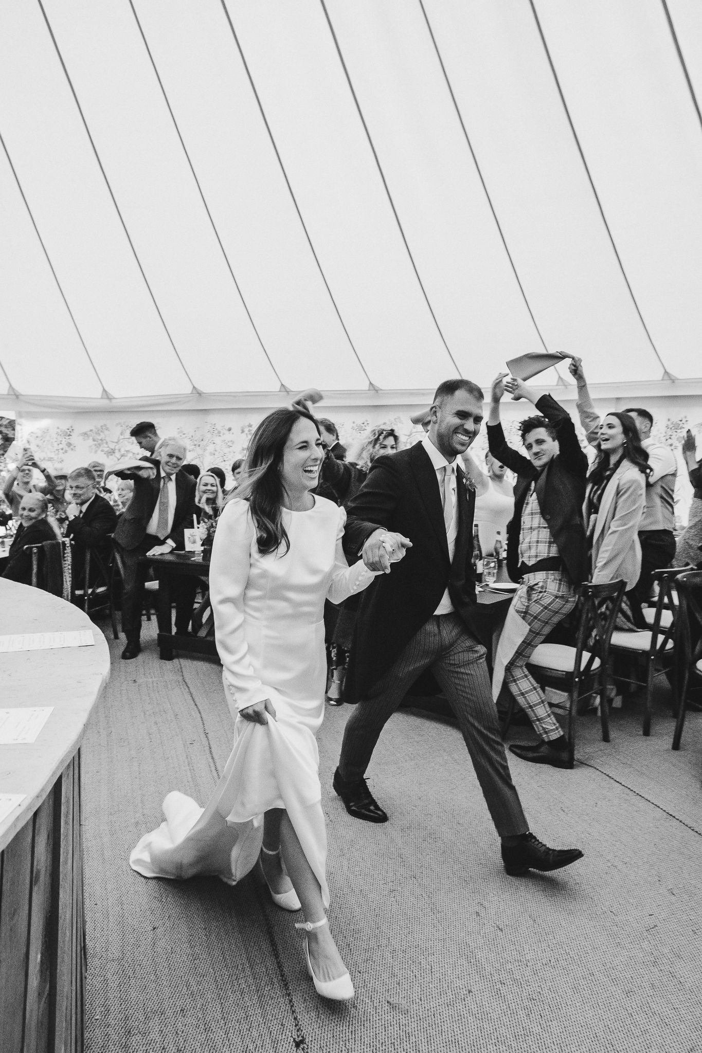 A couple dances energetically at a wedding reception inside a white tent while guests look on from their seats.