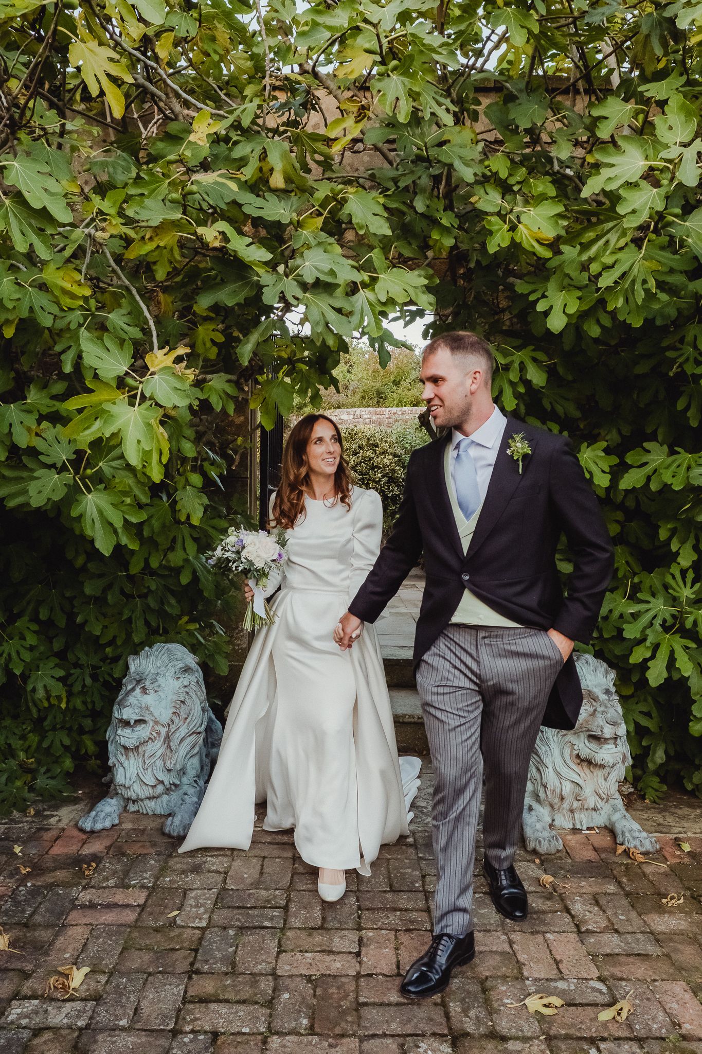 Newlyweds share romantic moments on brick patio with garden backdrop and stone lion statues.