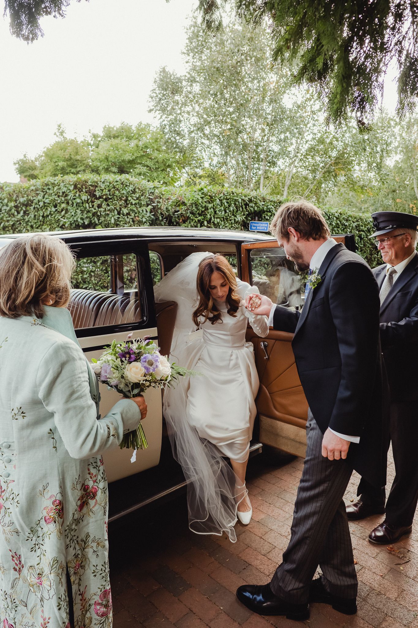 A bride in a white dress steps out of a vintage car with tan interior on a brick driveway during a wedding ceremony.