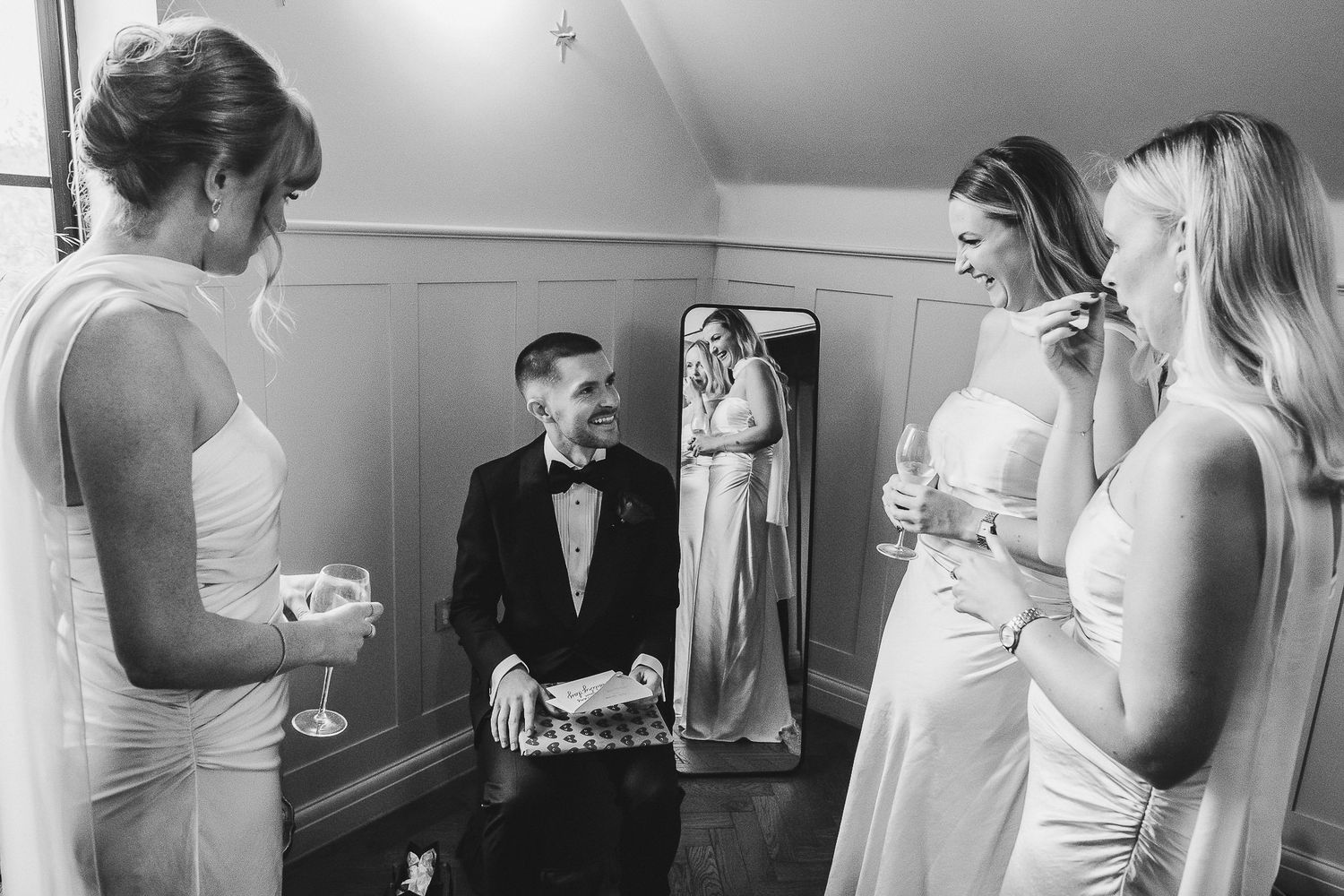 Black and white wedding photo showing bridesmaids in white dresses gathered around a mirror in a getting ready room.
