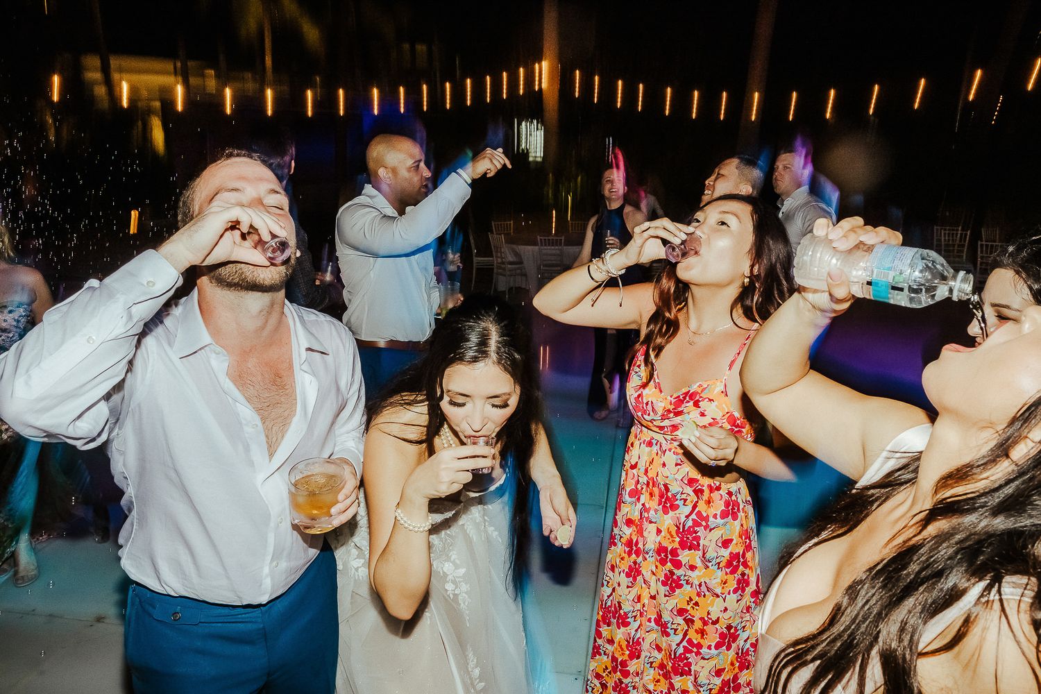 Party guests dancing and drinking at a nighttime outdoor celebration with string lights overhead.
