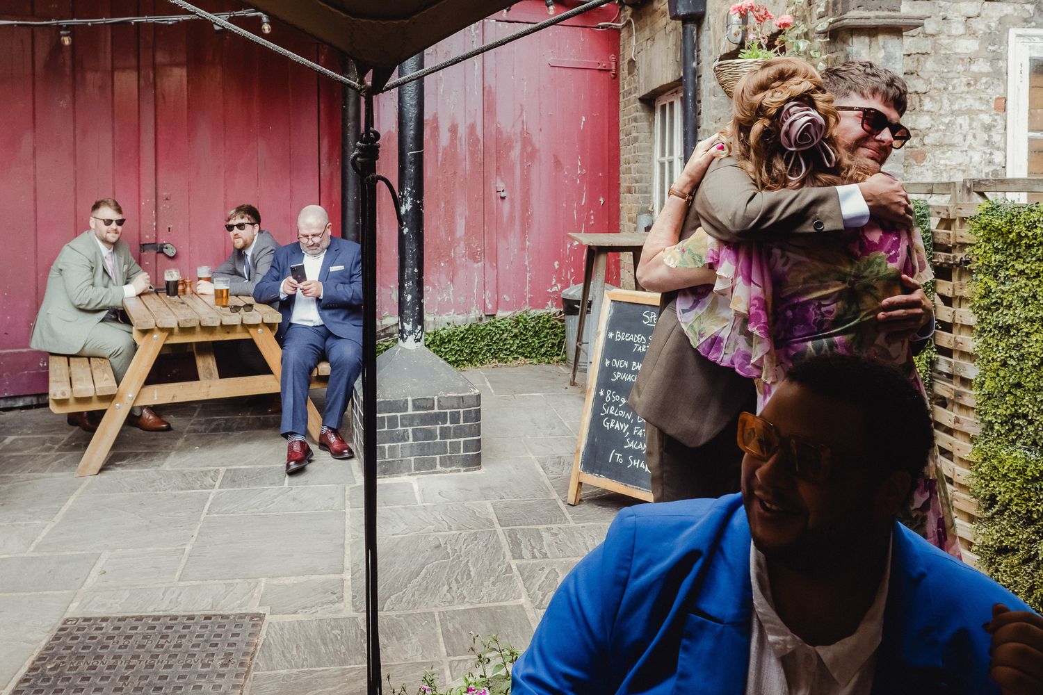 People gather at outdoor picnic tables near a rustic red barn wall during a social event.