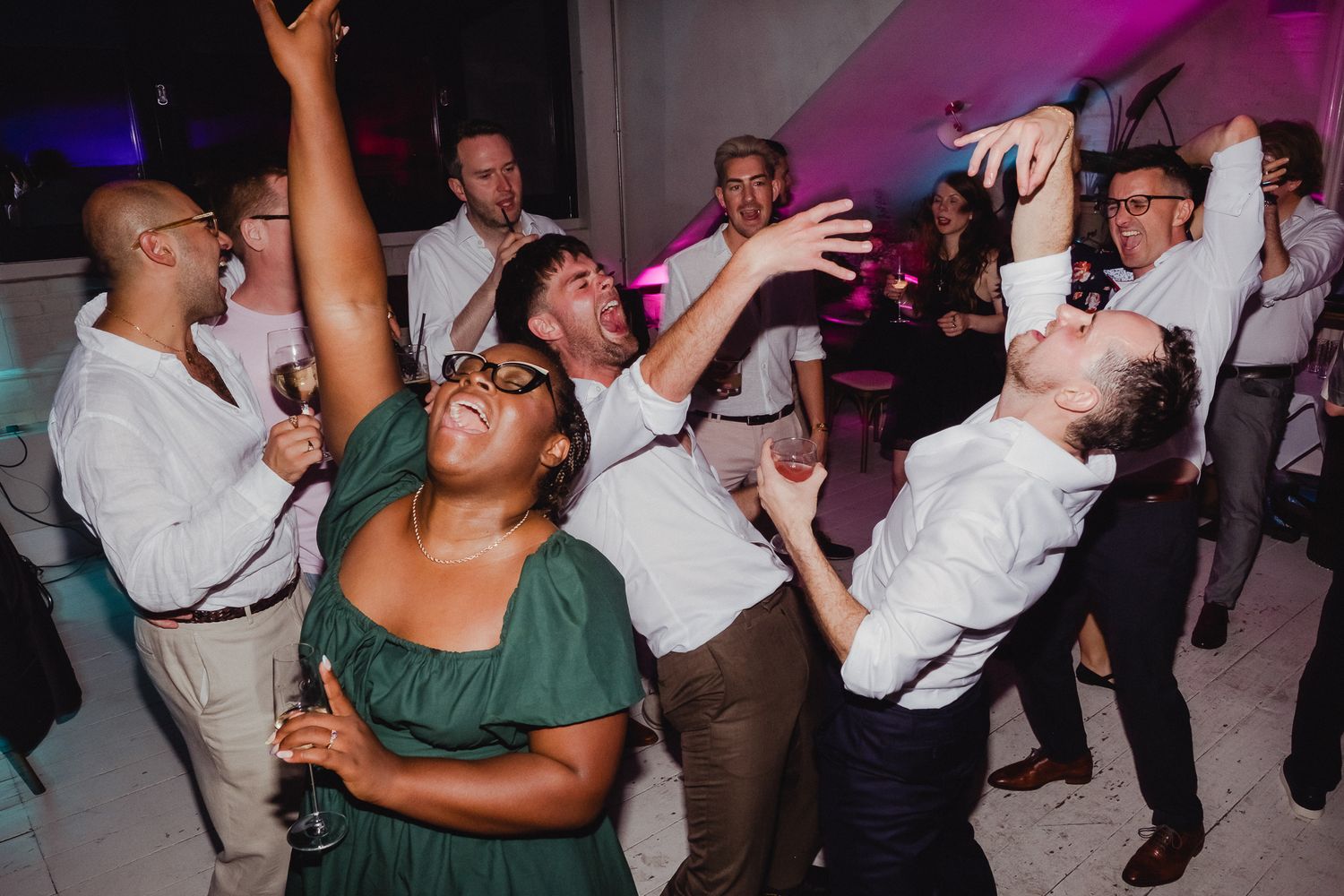 Wedding guests dance enthusiastically with raised arms on a purple-lit dance floor during a reception celebration.