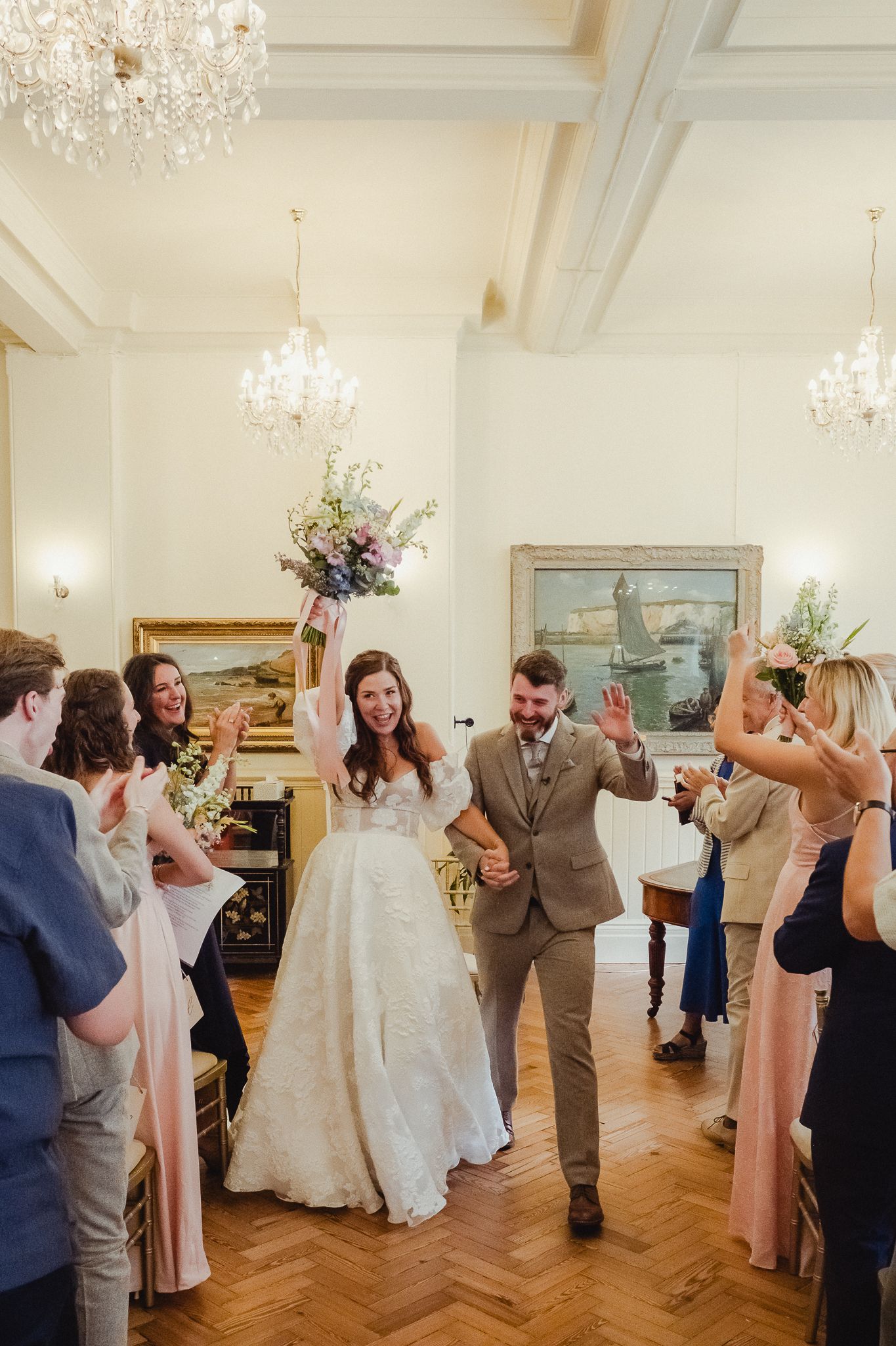 Wedding guests dance and celebrate together on a wooden dance floor under elegant chandeliers at an indoor reception venue.