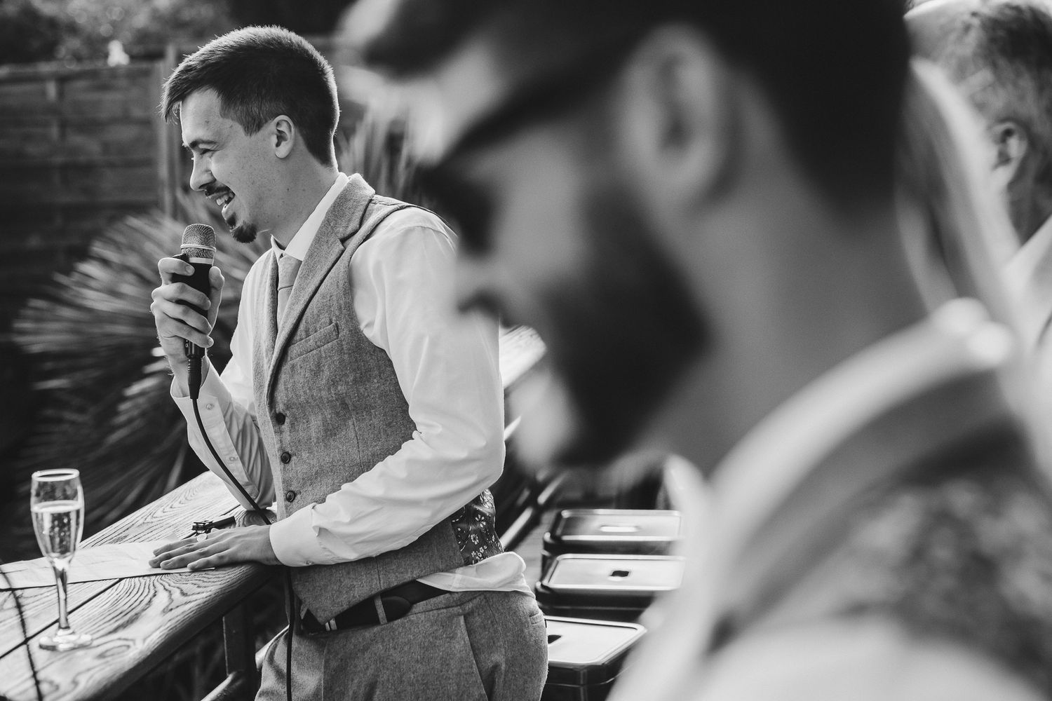 Black and white photo of someone in a vest and white shirt speaking emotively at a gathering with others seated nearby.