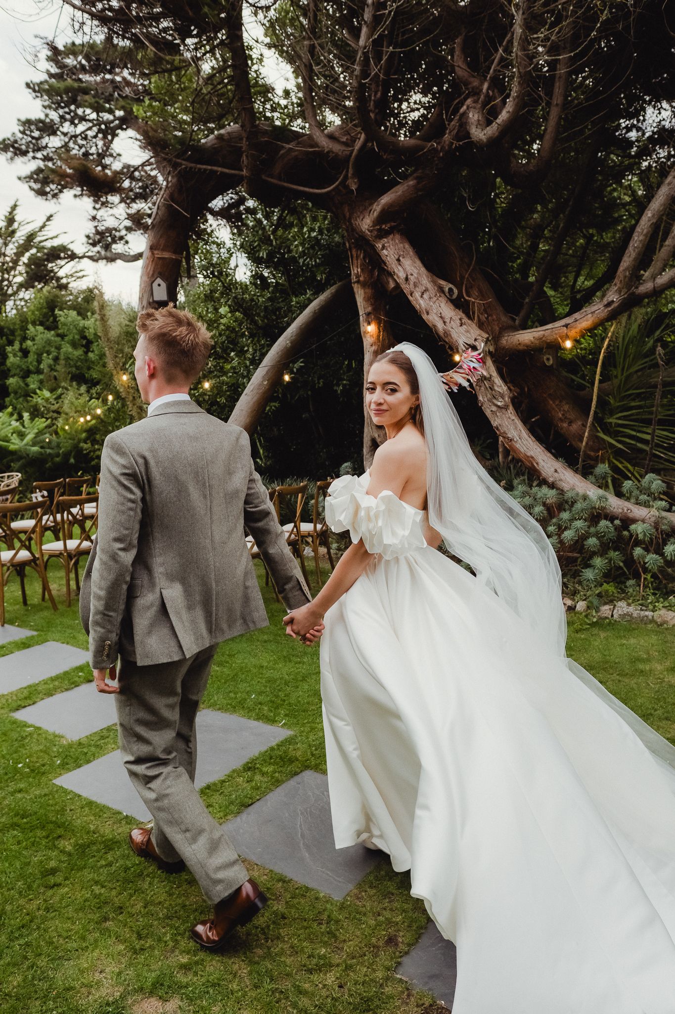 A couple walks through a garden setting with the bride's white veil flowing dramatically in the wind.