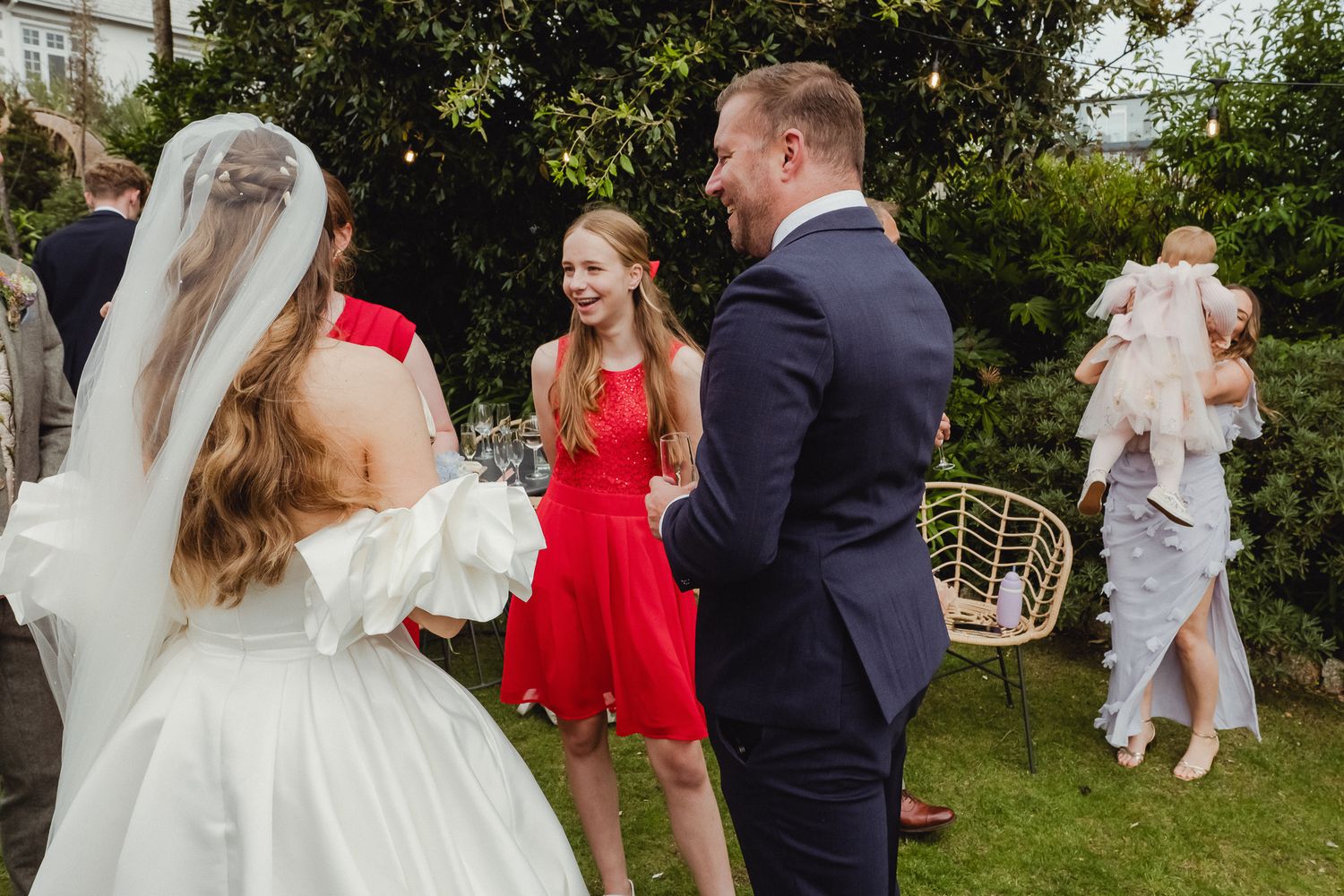 Wedding guests mingle in a garden setting with a bride in white dress and another guest in vibrant red dress.