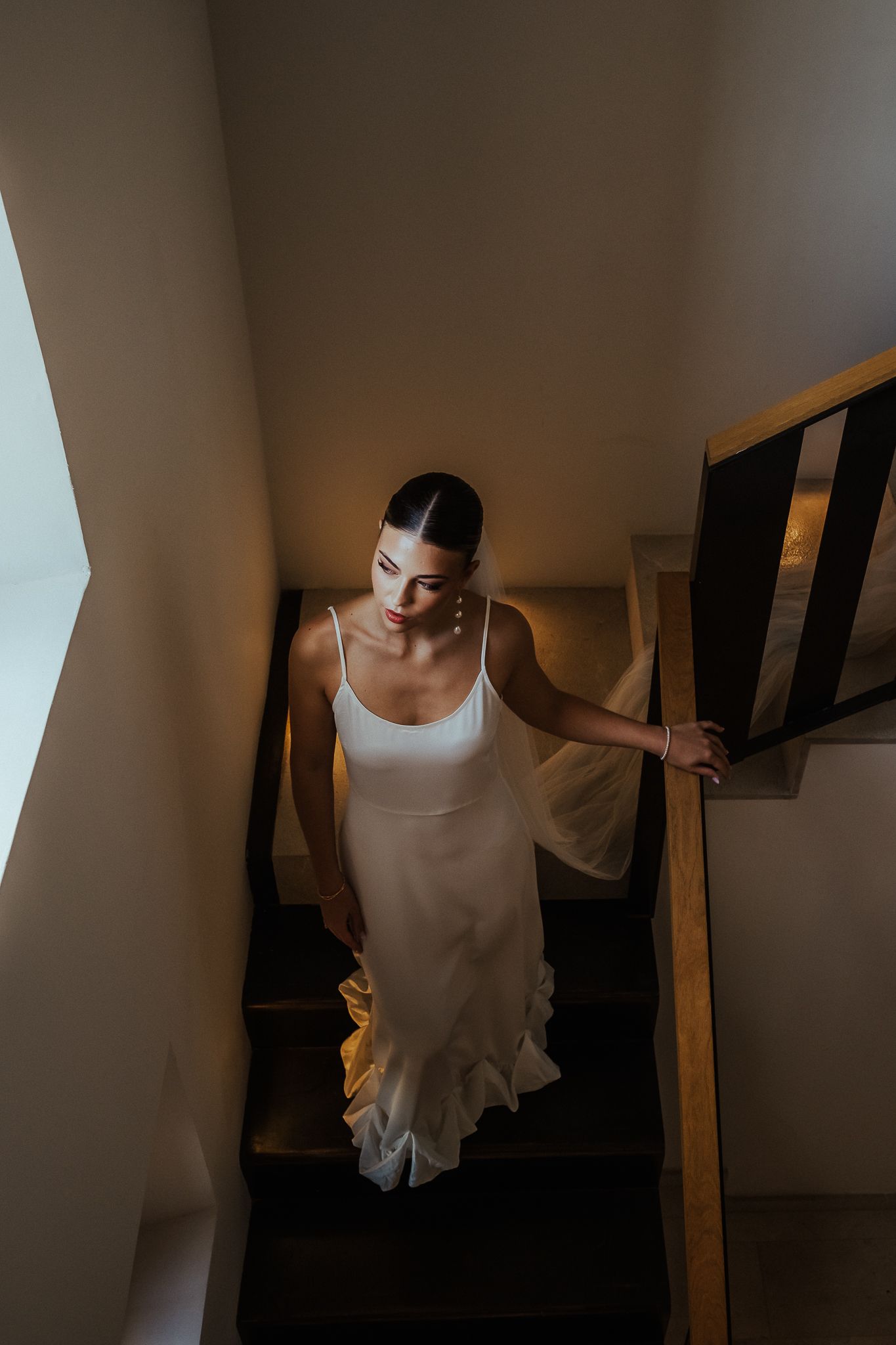 A person wearing a white sleeveless top stands in a dimly lit stairwell with wooden railings.