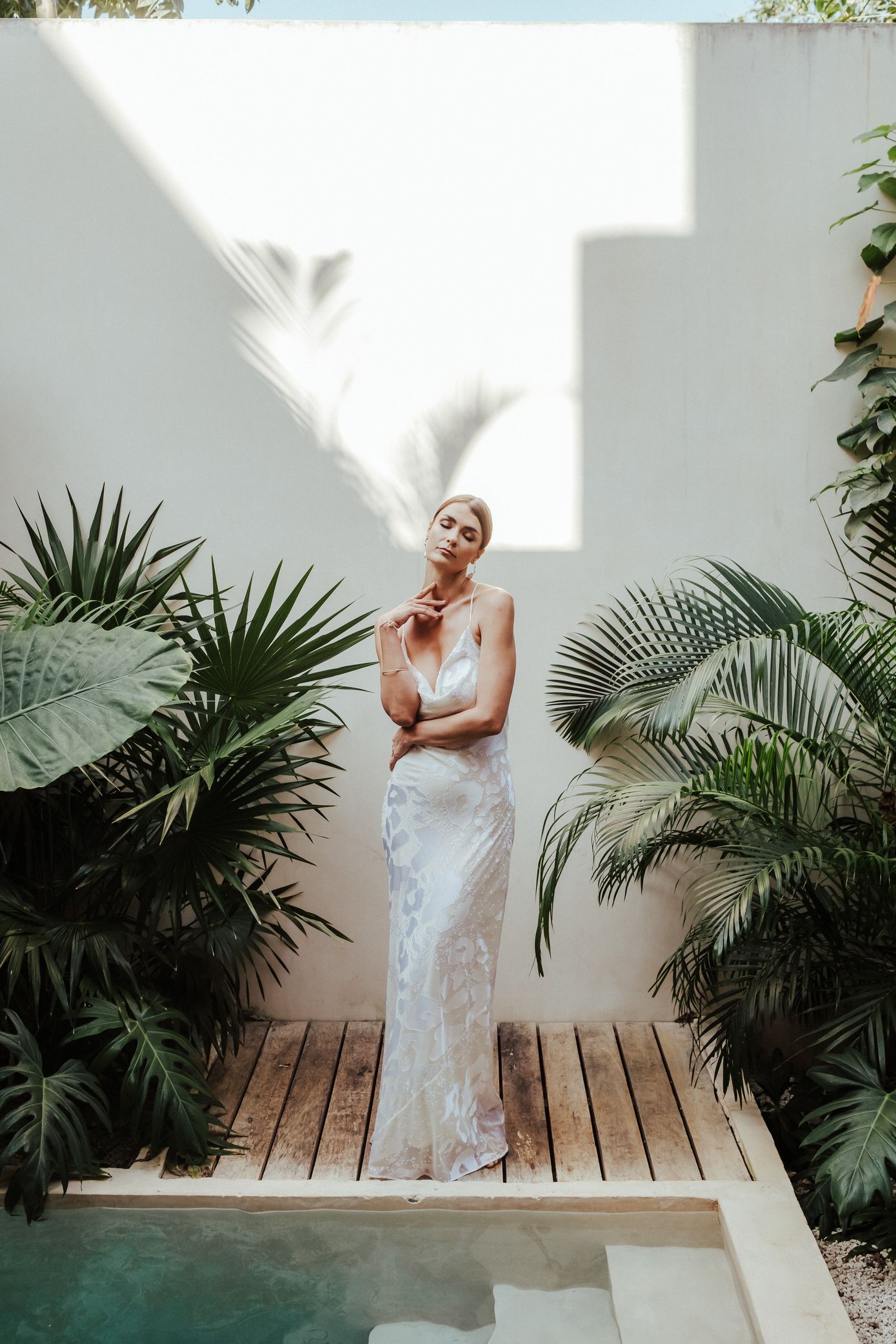 Elegant white lace gown photographed in a tropical courtyard setting with palm plants and wooden deck by a pool.
