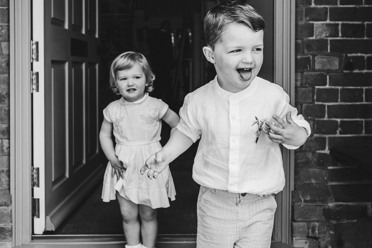 Black and white portrait of two children laughing on a doorstep in formal attire.