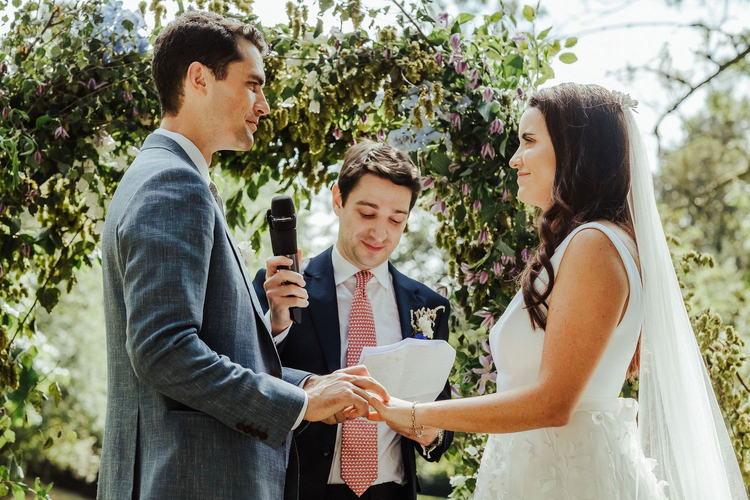 Wedding officiant conducts outdoor ceremony as couple holds hands in lush garden setting.