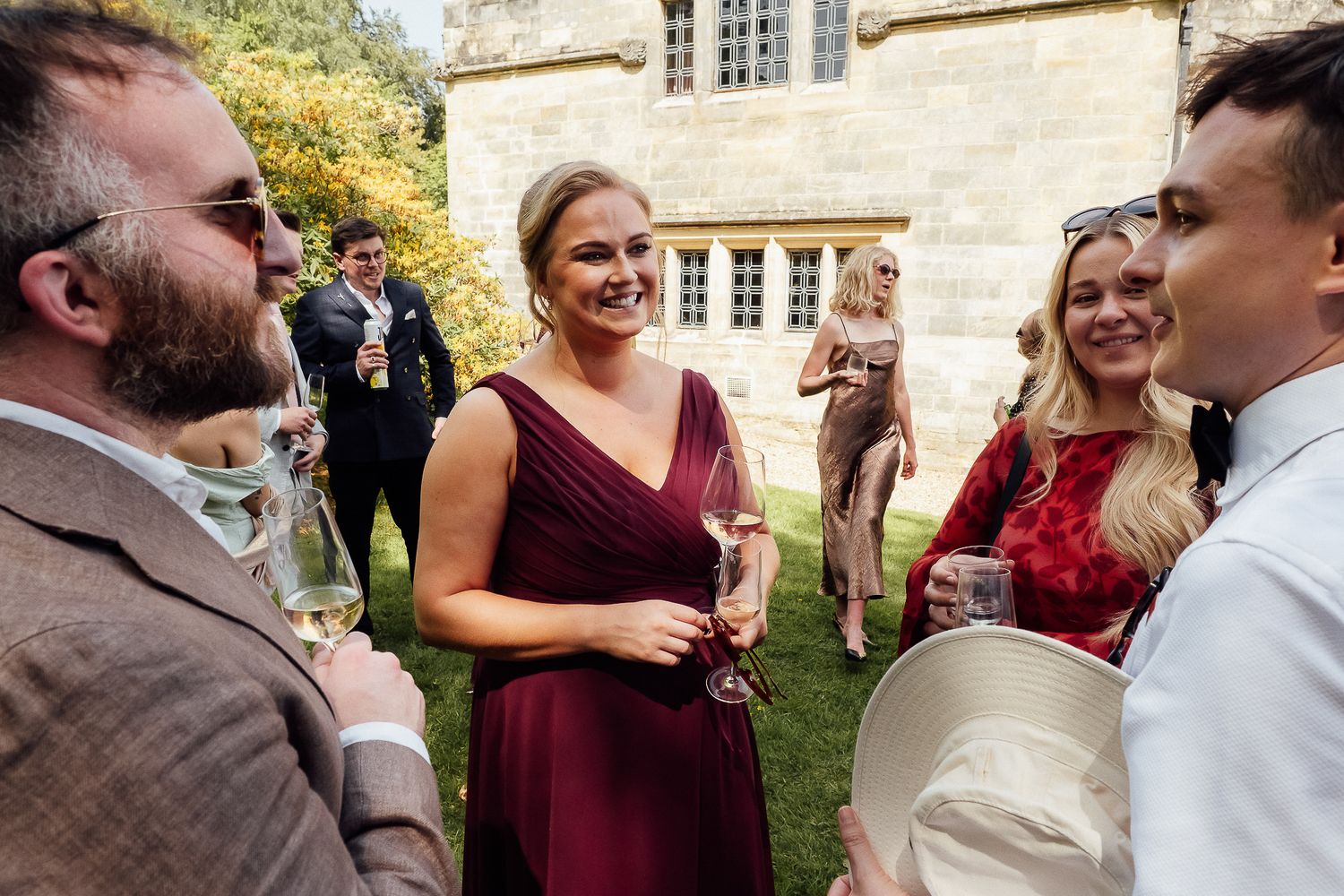 Wedding guests in formal attire socializing outdoors, holding champagne glasses near a historic stone building.