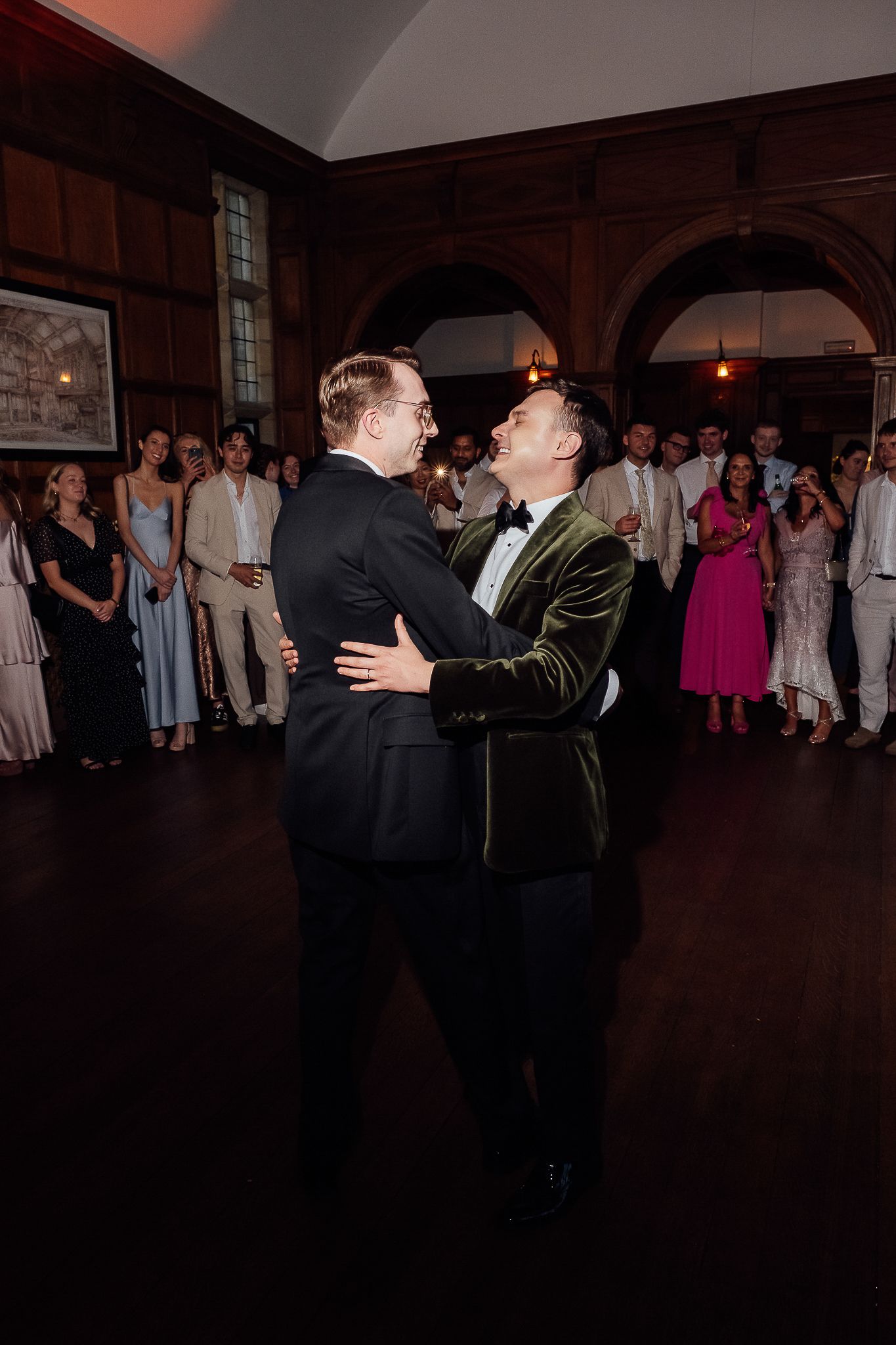 Two grooms share their first dance at a wedding reception in an elegant wood-paneled ballroom with guests watching.