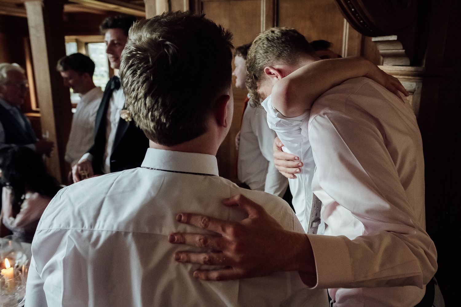 Two young men in white dress shirts share an emotional hug at a wedding reception.