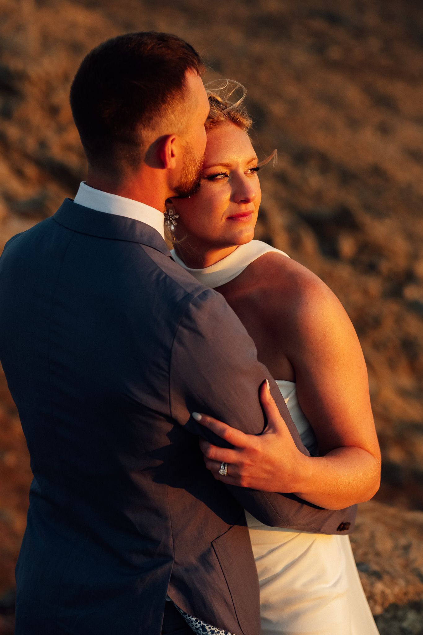 A bride and groom embrace at golden hour, bathed in warm sunset light against a rocky backdrop.