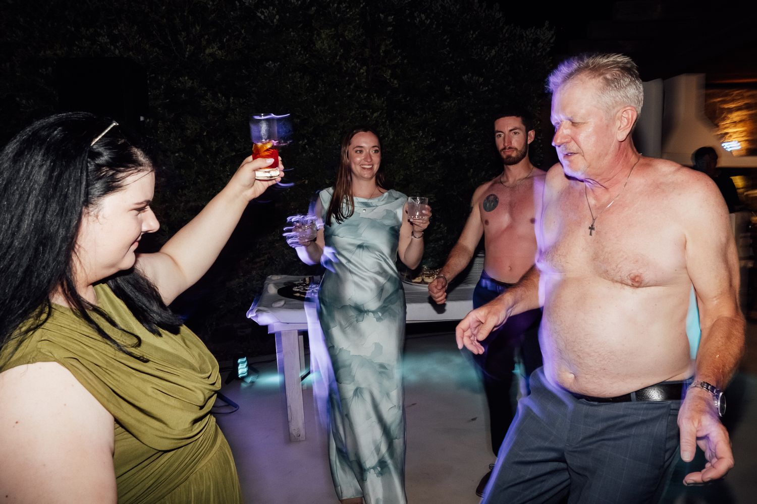 Group of adults dancing and celebrating outdoors at night, raising drinks and enjoying a lively party.