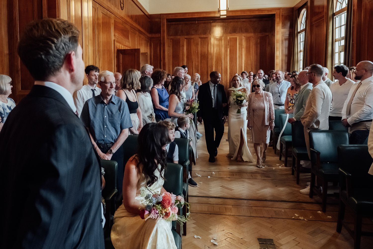 Bride and groom walk down the aisle between seated guests in a wood-paneled room during a wedding ceremony.